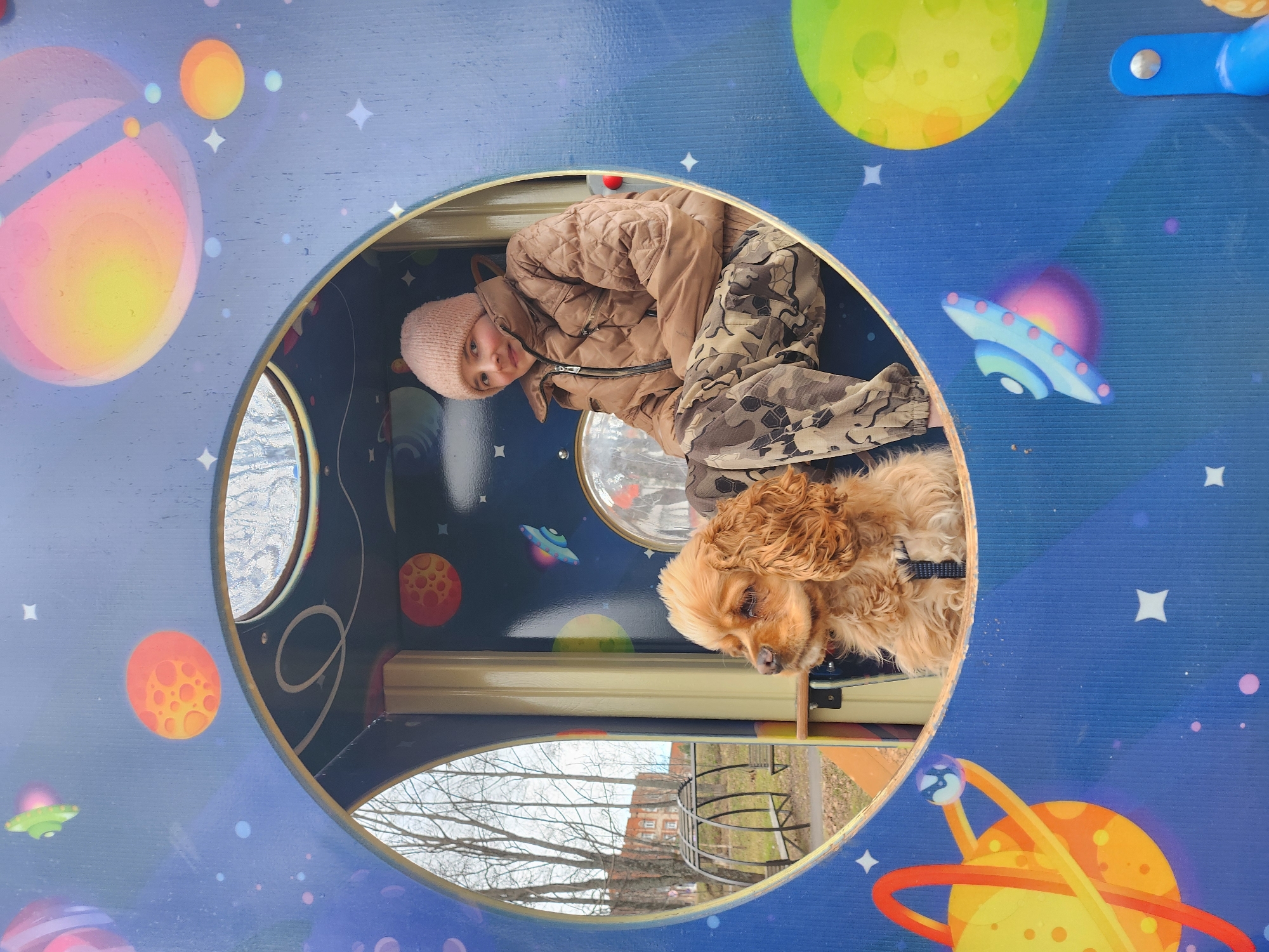 A young girl and a dog are captured inside a circular opening of a blue, space-themed play structure, likely in a park or playground in Krasnogorskoye, Russia. The image is taken during the daytime, under an overcast sky, suggested by the soft, diffused light and the bare branches of trees visible through openings in the structure.
The central subject is a young girl, possibly between 5 and 10 years old, who is looking directly at the viewer with a gentle, slight smile. She wears a light pink knitted beanie, a quilted light brown or tan jacket, and camouflage-patterned pants. Beside her, a medium-sized dog, possibly a Cocker Spaniel, with light brown, curly fur, is resting. The dog wears a black harness and is looking downwards.
The play structure itself is dark blue, decorated with whimsical illustrations of colorful planets (some with rings, some with craters), white four-pointed stars, and UFOs (one blue and pink, another green). The interior of the structure is also dark blue with similar space decorations. Through smaller window-like cutouts and a larger opening in the foreground, bare trees and a distant building can be seen, indicating an outdoor setting during a colder season like late autumn or early spring. No visible text is present in the image.