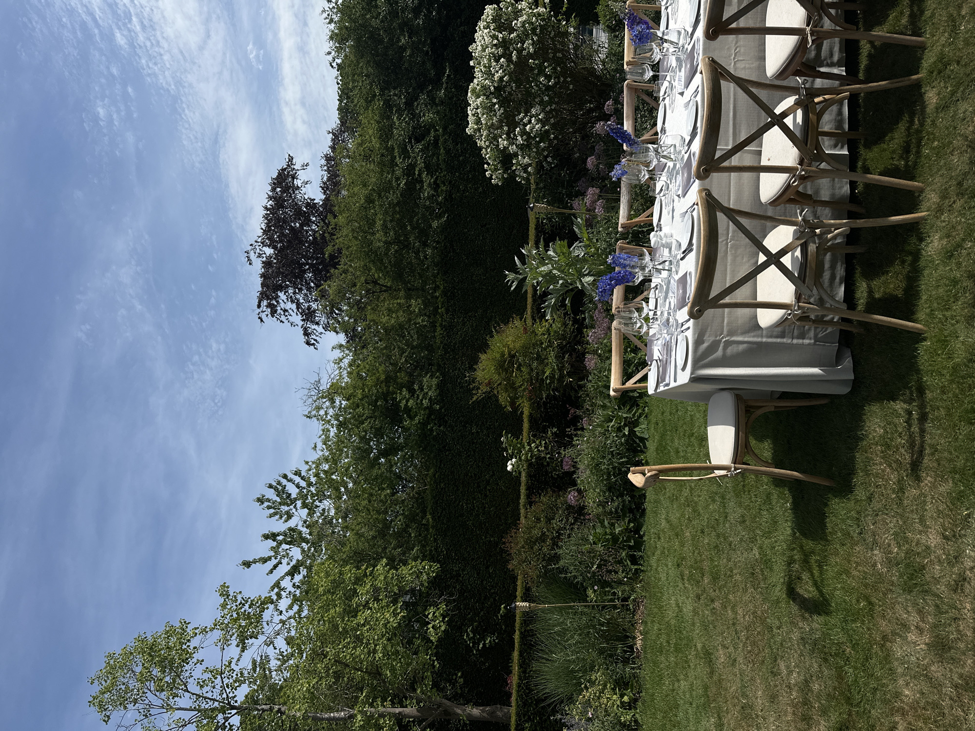 The image depicts an outdoor dining setup in a garden setting, likely in the city of Cambridge, United Kingdom. A long, grey-clothed table is arranged with place settings, including plates, glasses, and cutlery. Wooden chairs with off-white cushions are placed around the table. Small arrangements of blue flowers decorate the table's center. The dining area is situated on a lawn, bordered by various trees, hedges, and flowering plants. The sky is partly cloudy.