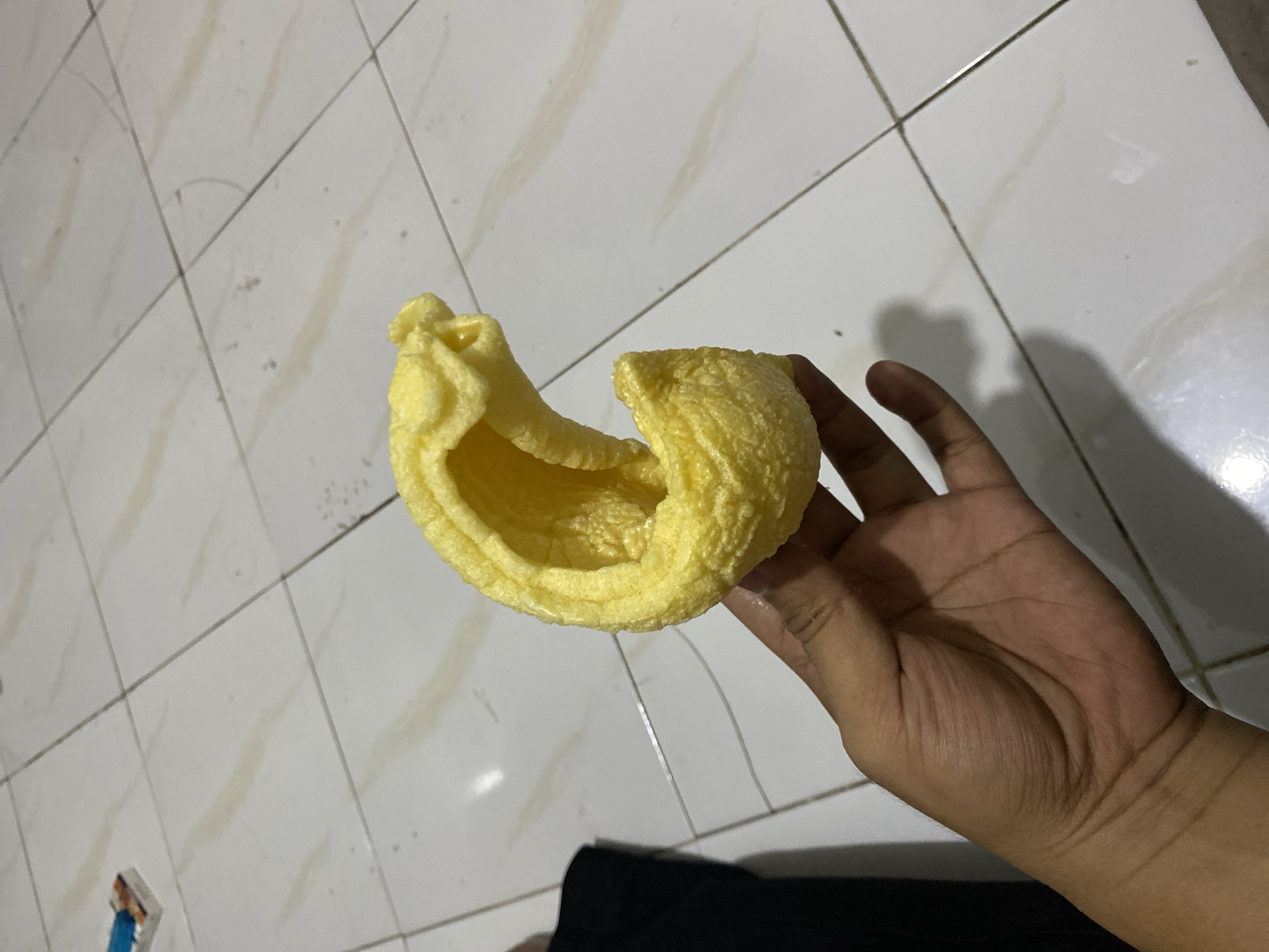 A hand holds a puffed, bright yellow snack that is curved into a crescent shape. The snack has a textured surface and appears hollow. The hand, with visible skin folds and lines, is positioned in the lower portion of the image. The background consists of a light-colored tiled floor with faint, irregular lines of a darker hue. A dark, soft surface, possibly fabric, is visible in the lower left corner, indicating a person's clothing. The overall lighting suggests an indoor environment with a shadow cast by the snack and hand onto the floor. The location is Tebel, Indonesia.