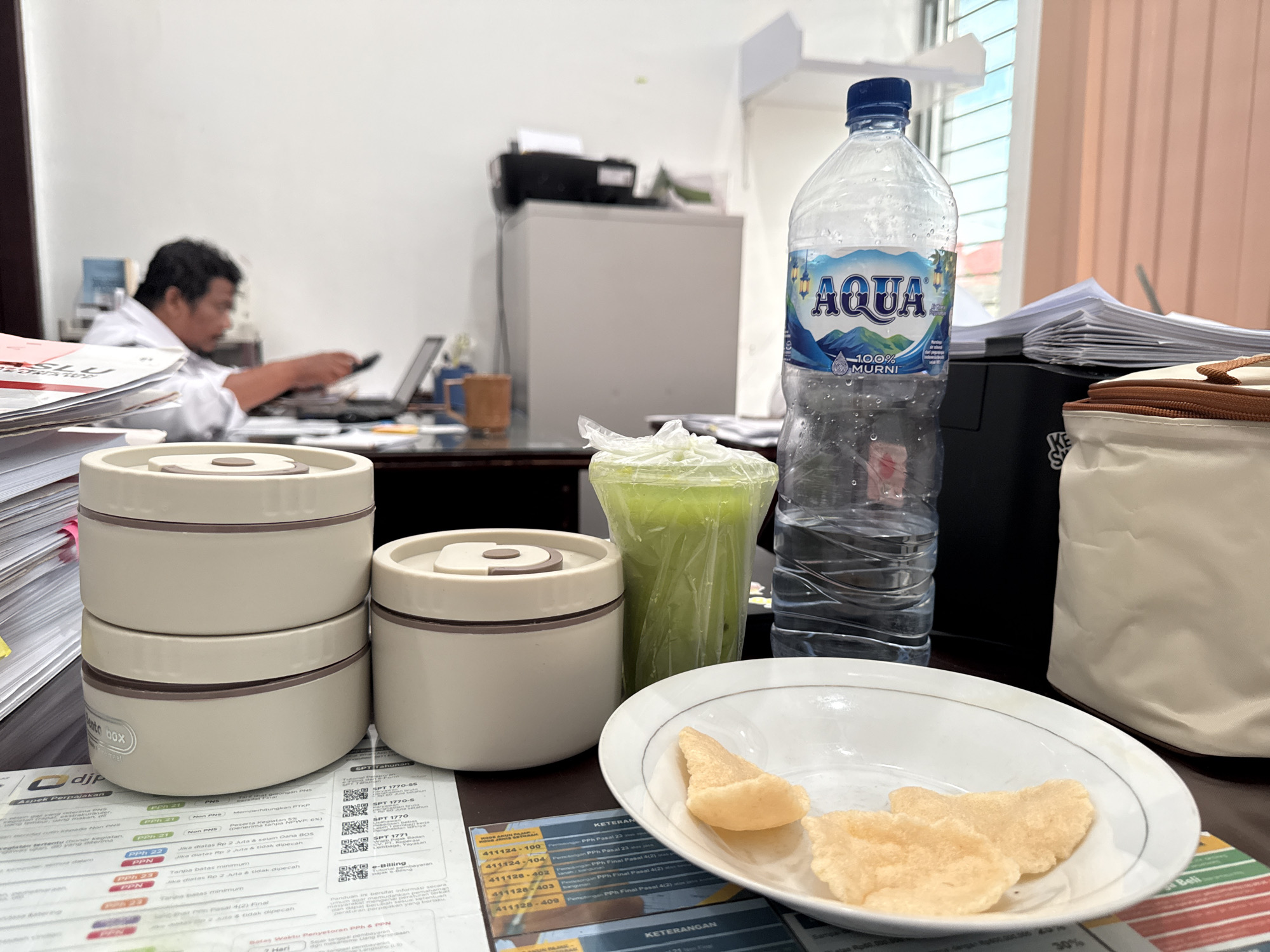 The image shows an office desk with a man working on a laptop in the background. In the foreground, there is a stack of documents, two stackable lunch boxes, a cup of green liquid in a plastic bag, a bottle of "AQUA" brand mineral water, a plate with crackers, and a small cooler bag. Some of the documents on the desk have text in Indonesian, including the words "SPT Tahunan," "PPh 21," "PPN," and "KETERANGAN." Based on the text visible in the image, the documents appear to be related to tax matters.
The office setting and the presence of tax-related documents suggest the location could be in Lhokseumawe, Indonesia. It is only a suggestion.
