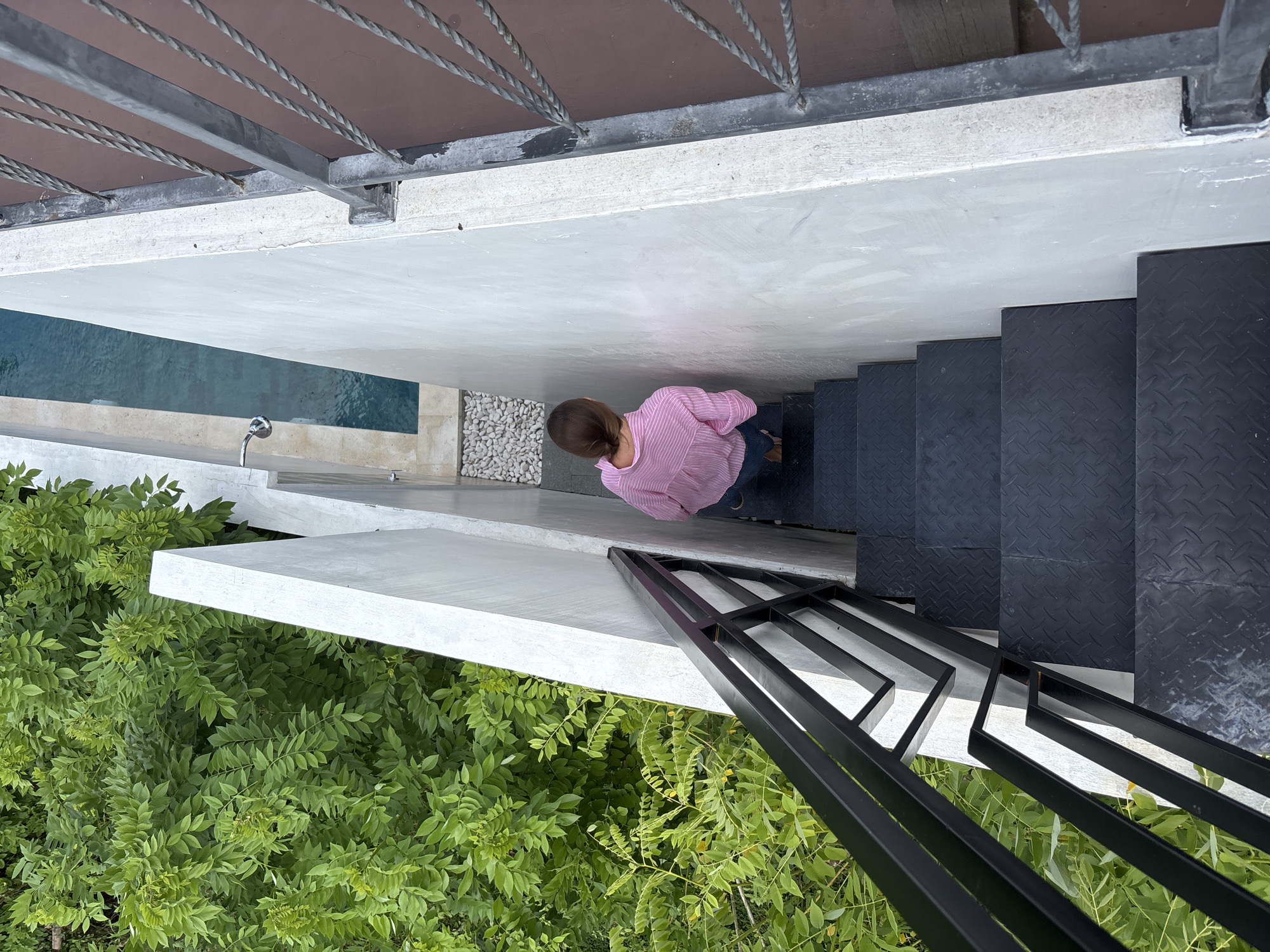 Captured from an elevated perspective, the image reveals an outdoor architectural space in Ungasan, Indonesia. A woman with short brown hair, dressed in a pink and white striped shirt and dark pants, stands on the landing between a set of black metal stairs and a white concrete platform.
The staircase descends to a lower level, bordered by a black metal railing. Lush green foliage is visible below. Adjacent to the landing is a shallow pool of water with white decorative stones and an outdoor shower fixture. The pool is bordered by a white concrete surface.
Overhead, a brown structure with a metal railing provides shade. The scene is characterized by the stark contrast between the cool tones of the concrete and metal, and the natural warmth of the vegetation and wood.