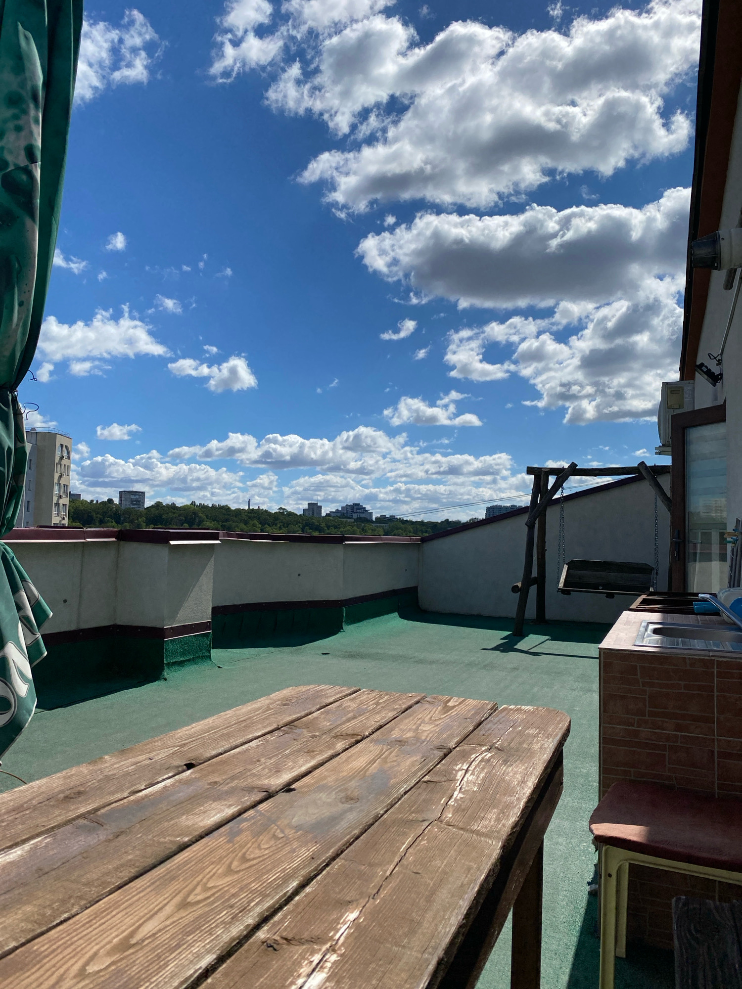 The image depicts a rooftop terrace on a sunny day with a bright blue sky dotted with white, fluffy clouds. In the foreground, a weathered wooden picnic table occupies the lower portion of the frame. The terrace floor is covered in a green synthetic material.
Further back on the terrace, a rustic wooden swing set stands, with a dark-colored seat hanging from chains. To the right, an outdoor kitchen counter with a sink is visible, adjacent to a brick-like tiled base and a red stool.
Beyond the terrace railing, a cityscape unfolds. Buildings of various sizes, including a tall apartment building, are scattered amongst a lush green treeline. In the distance, more buildings and hints of urban infrastructure are discernible. A partial view of a green patterned awning or curtain is visible on the left side of the image. The overall impression is one of an urban outdoor recreational space. Based on visual cues, the location is inferred to be Chișinău, Moldova.