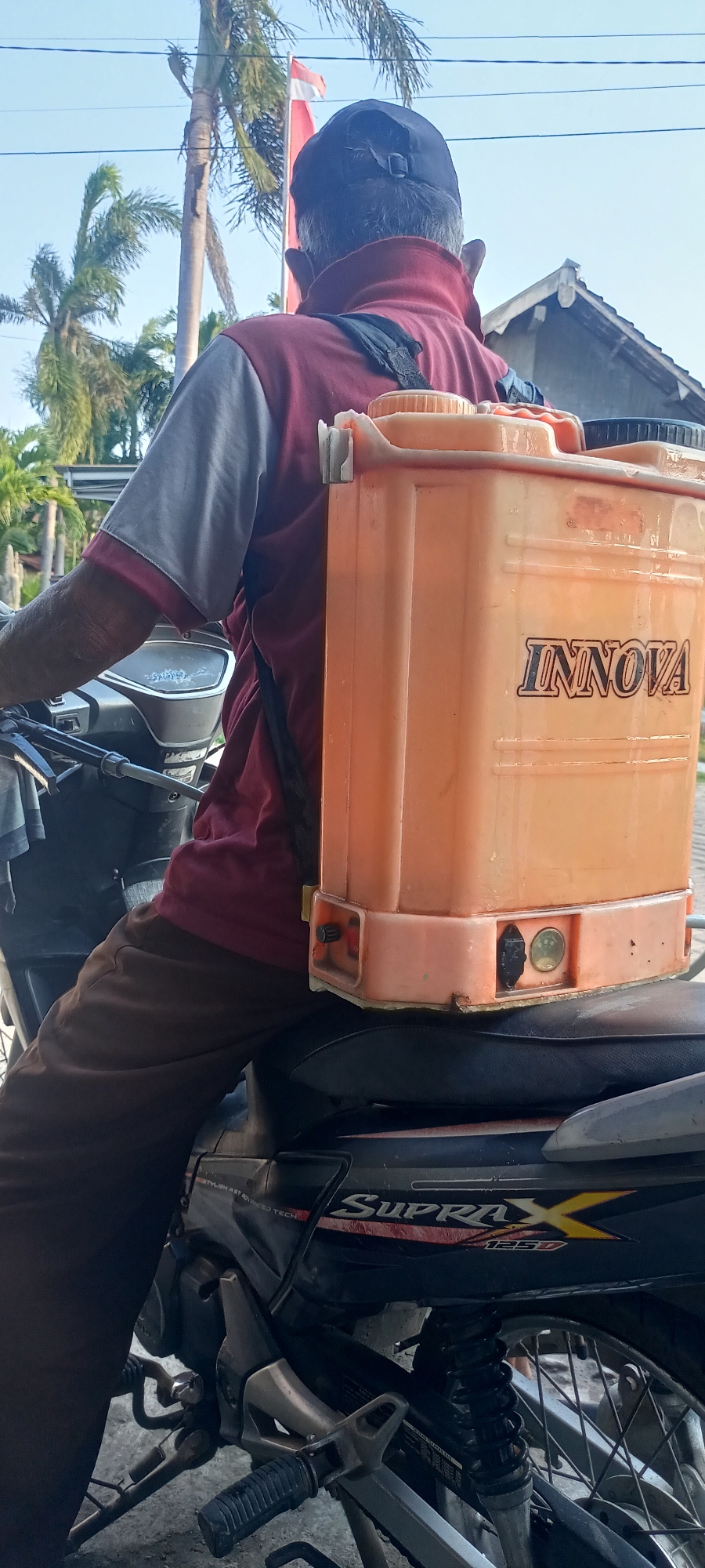 In Gempolklutuk, Indonesia, a person is seated on a motorcycle, facing away from the camera. They are wearing a maroon and grey shirt, brown pants, and a black baseball cap. Strapped to their back is a large, orange sprayer tank with the brand name "INNOVA" visible in black lettering. The motorcycle, a Honda Supra X 125, is dark in color, with some decals on the side panel. The background features palm trees, a house with a dark roof, and a bright blue sky, suggesting an outdoor, possibly rural or suburban, setting during daylight hours. The overall scene depicts an individual preparing for or in the process of using the sprayer, possibly for agricultural or pest control purposes.