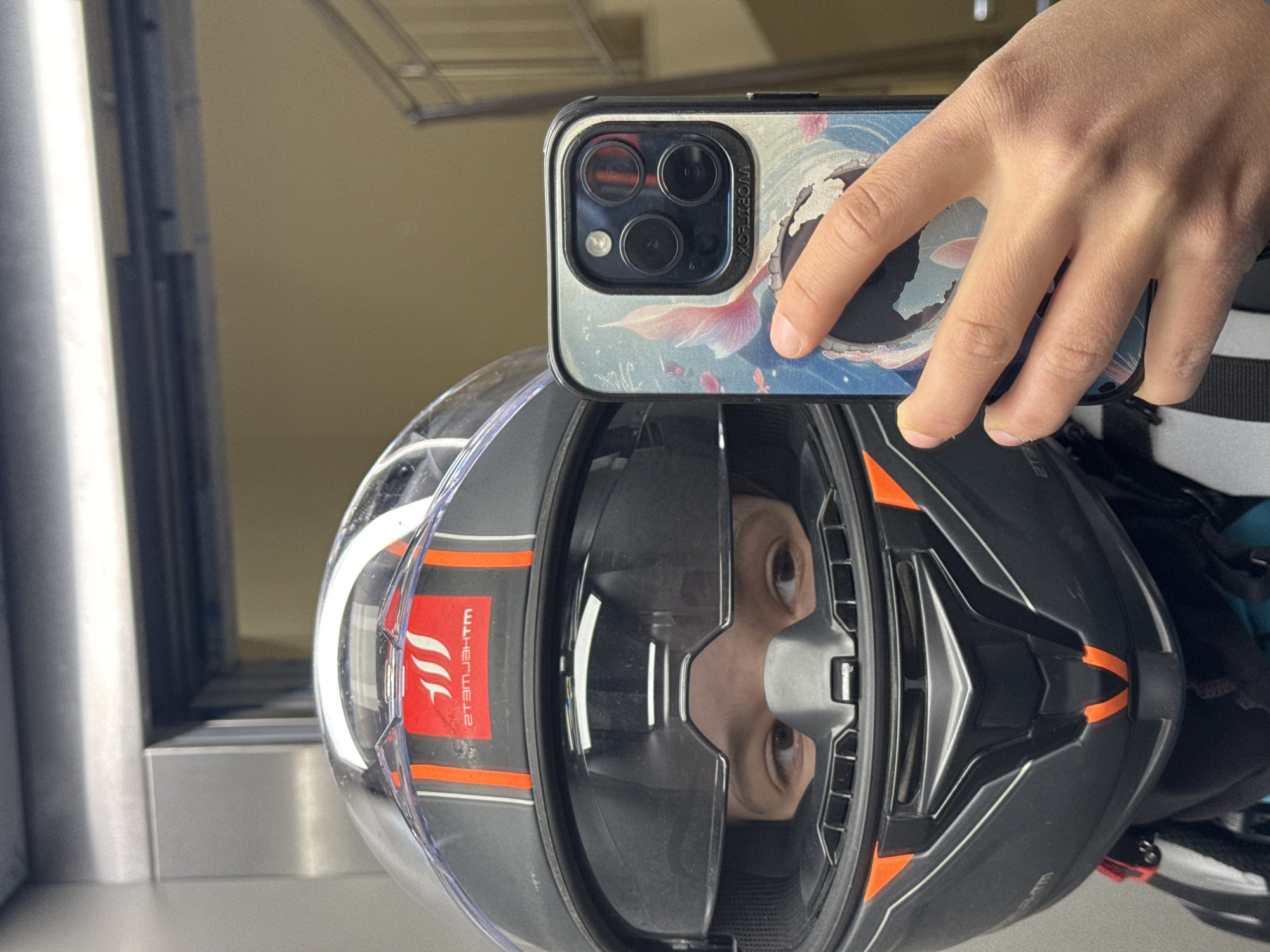 A person is taking a selfie in a mirror or reflective surface, holding an iPhone with a patterned case. The reflection shows the person wearing a gray and black motorcycle helmet with orange accents. The helmet obscures most of the face, but the eyes are visible in the reflection. The background is blurry, but there appears to be a metal structure and a light-colored wall. The location in the city of Varna, Bulgaria cannot be determined from the image.
