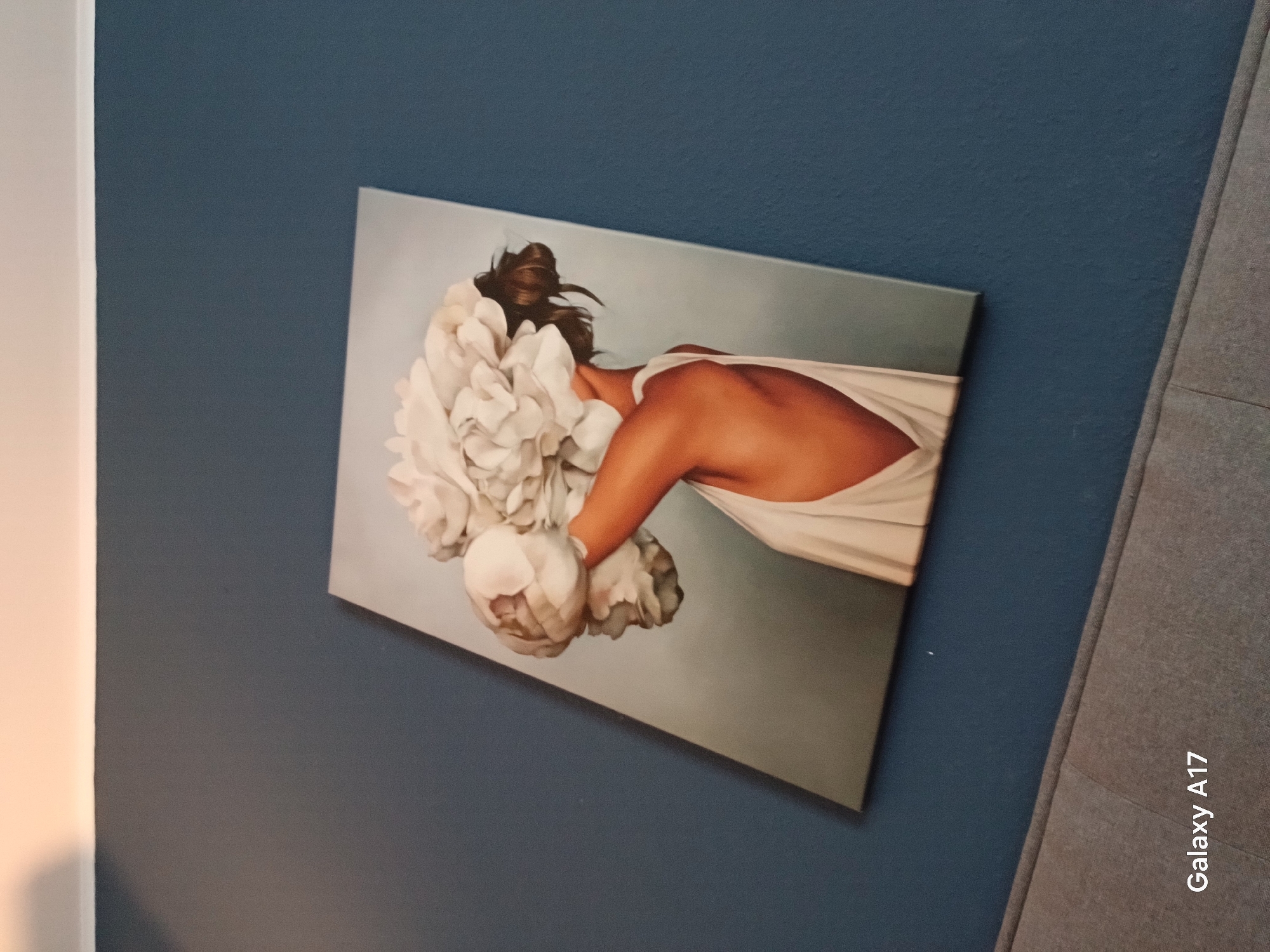 The image shows a painting hanging on a blue wall, adjacent to a gray fabric-covered surface. The painting depicts a woman, viewed from the back, with white flowers covering part of her face and shoulder. Her bare back and part of her arm are visible, and she is draped in a white cloth. The background of the painting is a neutral gray. The photo was taken with a Galaxy A17. There is no location in Yekaterinburg, Russia, given in the image.
