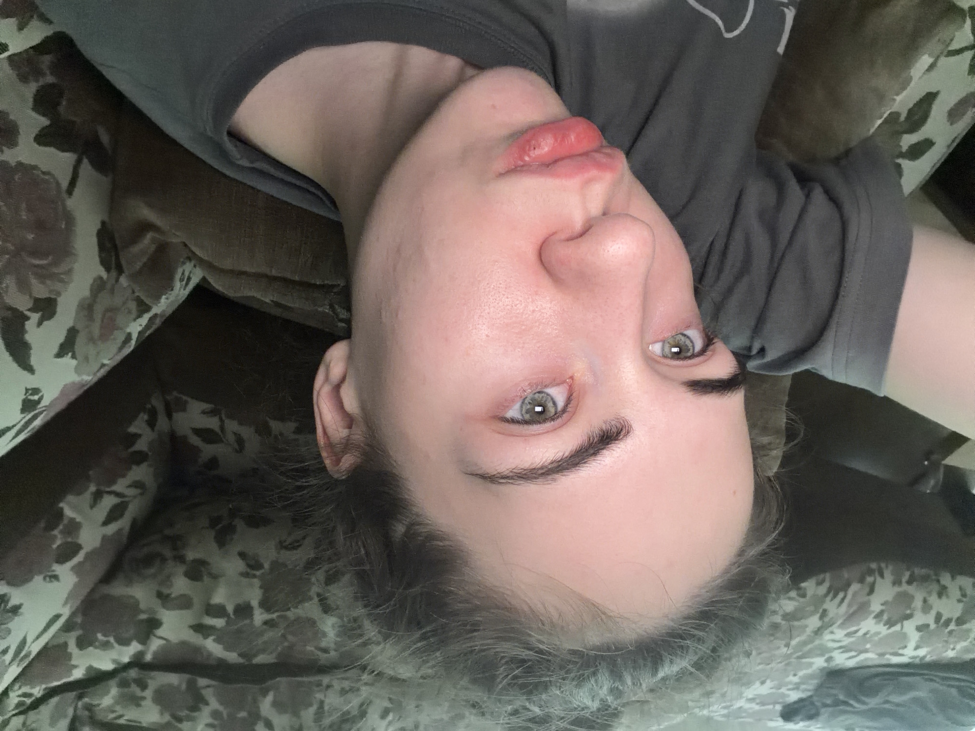 The image shows a close-up, high-angle view of a young Caucasian female lying on her side. Her face is the primary focus, with her lips pursed. She is wearing a dark gray t-shirt. The background consists of floral-patterned upholstery and a brown textured surface, suggesting she is on a couch or bed. There are no discernible elements in the image that would indicate a specific location within Zavodoukovsk, Russia.