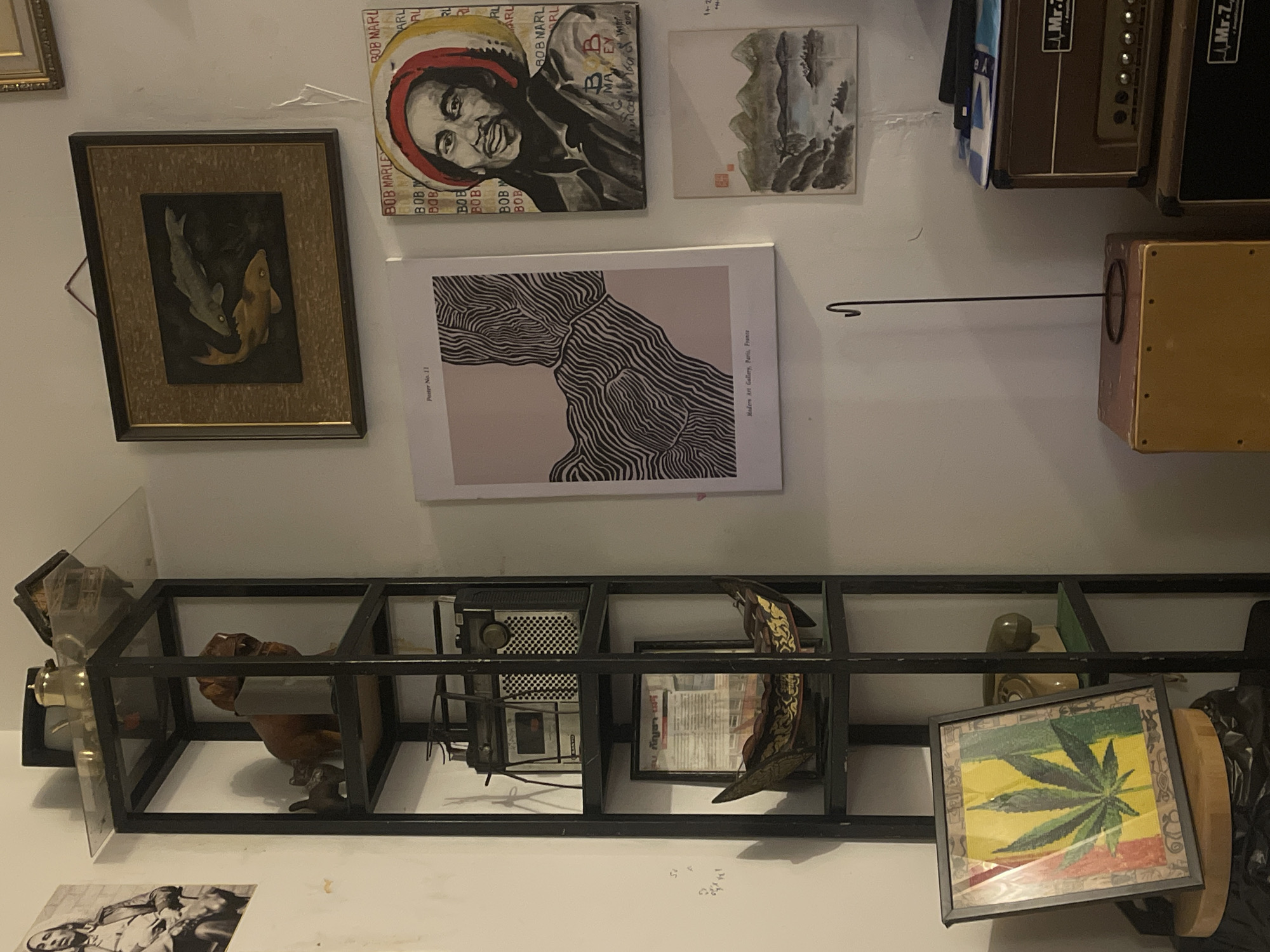 The image depicts an indoor scene, possibly a room in Chalong, Thailand, filled with various decorative items and musical equipment. A tall, black metal shelving unit stands prominently on the left, displaying a collection of objects. The top shelf holds a small building model and a metal teapot. Below that, a wooden carving of a dog sits next to a smaller animal figurine. Further down, a vintage radio and a framed document are visible. At the bottom of the shelf unit, a decorative boat-like object with intricate patterns rests.
To the right of the shelving unit, the wall is adorned with several framed artworks. A framed painting of two fish hangs high on the wall. Below it, a portrait of Bob Marley is displayed, surrounded by repeated text. Another smaller framed painting depicts a mountainous landscape with a body of water. To the left of the Bob Marley portrait is a poster with abstract black and white lines.
In the foreground on the left, a framed artwork featuring a cannabis leaf with a red, yellow, and green background is leaning against a circular object, likely a drum, which is placed next to a trash can. On the right side of the image, two stacked amplifier cabinets are visible, with labels indicating "Mr.Z." and "Amplification." A thin, black metal stand with a hook extends upwards from a wooden box. The lighting in the room is dim, suggesting it may be evening or the curtains are drawn. The overall impression is a personal space decorated with a mix of cultural and artistic elements.