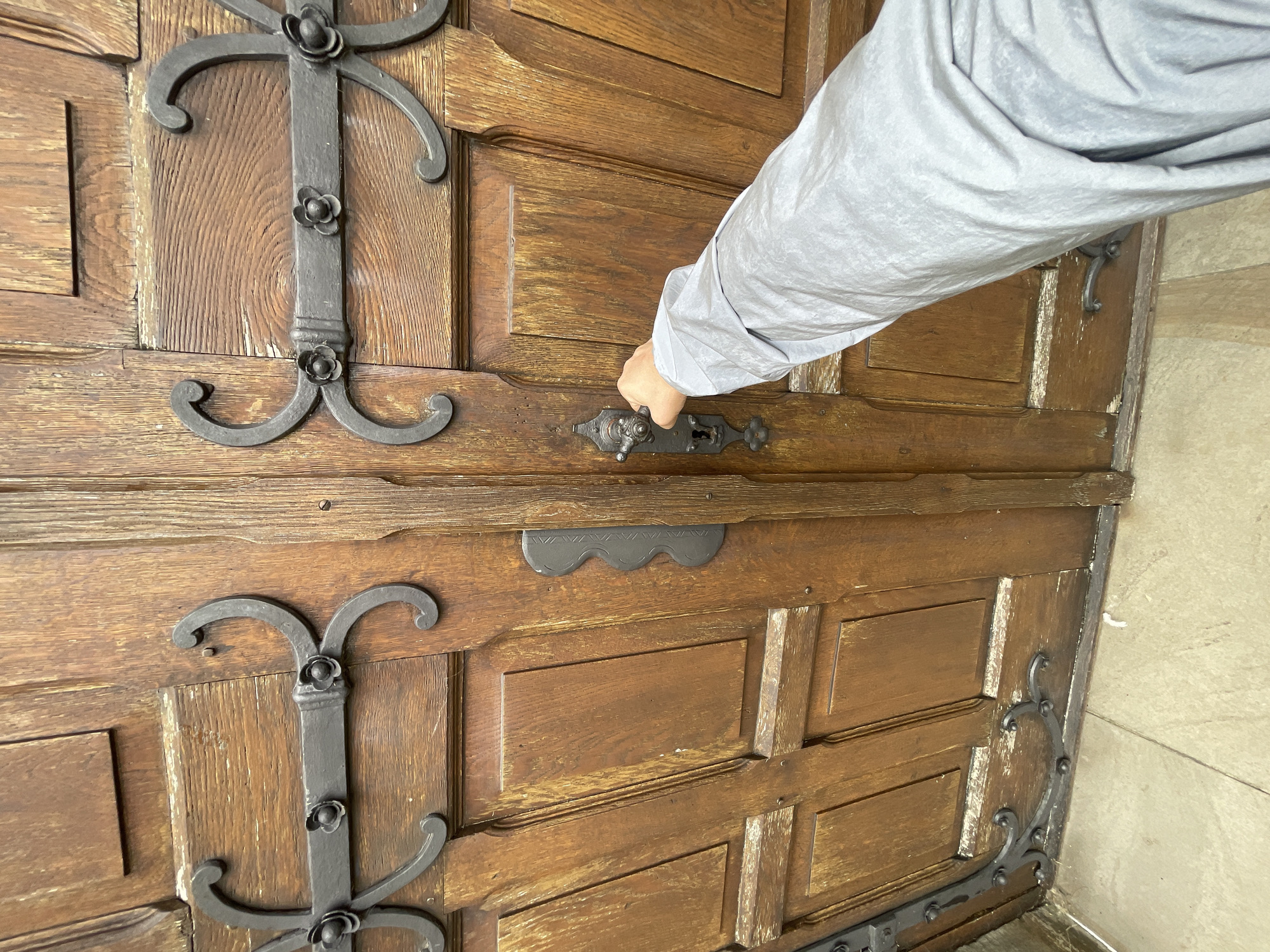 A person's hand, clad in a light grey sleeve, is reaching out to turn the knob of a large, ornate wooden door. The door is constructed from thick, weathered planks and features intricate dark metal hardware, including decorative straps with flower motifs and a prominent, old-fashioned lock mechanism. The door appears to be part of an entrance to a building with a historical or traditional architectural style. The surrounding environment suggests an outdoor setting, with light-colored stone tiles visible at the bottom of the frame, indicating a pathway or entryway. The image captures a moment of entry or exit, with the hand poised to operate the door. This scene is located at the historical St. John's Church (St. Johannes der Täufer) in Salzkotten, Germany.