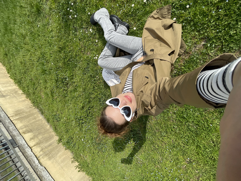 A female individual is depicted lying on her back on a patch of green grass under direct sunlight. She is positioned diagonally in the frame, facing the camera with a slight smile. The individual has light brown hair pulled back and is wearing white, heart-shaped sunglasses. Her attire consists of a brown/khaki trench coat, a black and white striped top, light grey jeans, and black shoes. Her legs are bent, with her left foot pointed towards the right. Her right arm is extended into the foreground, holding a device (likely a smartphone) to capture the image, revealing a black and white striped sleeve under the trench coat cuff. A clear shadow from her head and arm is cast on the grass to her left. The grass is interspersed with numerous small white flowers. In the background, an angled concrete retaining wall, light in color, is visible, topped by a dark metal fence or railing. The precise location within Budva, Montenegro, is not discernible from the visual information provided.