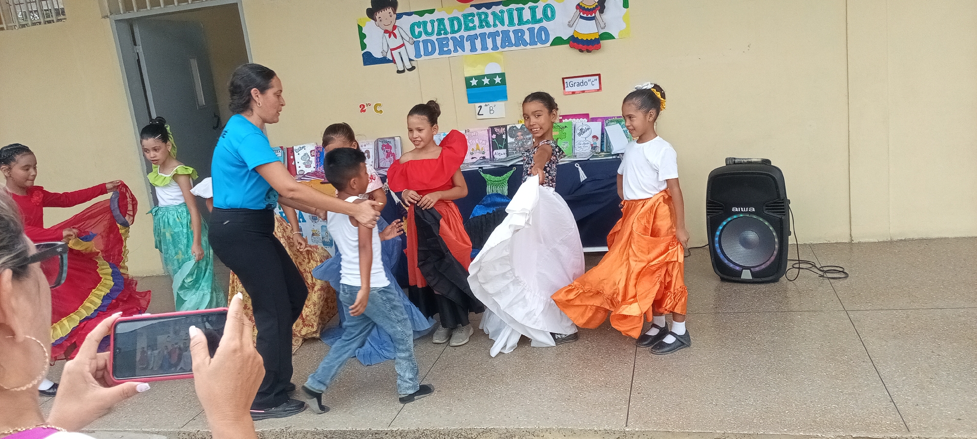 The image shows a group of children and an adult woman outdoors, likely at a school or community event. The children are dressed in traditional-style clothing, suggesting a cultural performance or celebration. The woman is likely directing or assisting them. A person in the foreground is using a smartphone to record the scene. A portable speaker sits on the right side of the image. The backdrop includes a banner or display with text and illustrations.