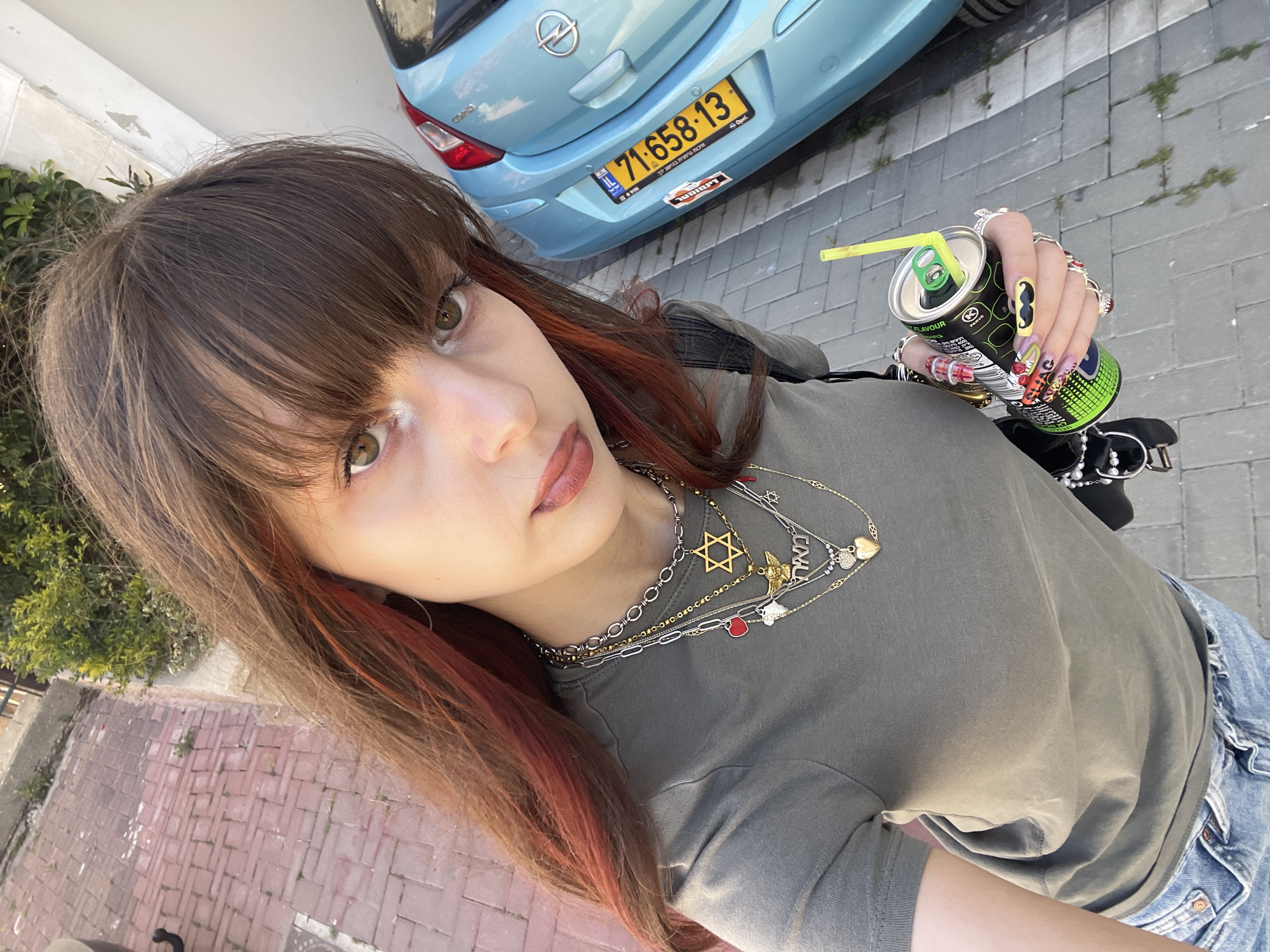 A young woman is shown in a high-angle selfie, holding a green and black energy drink can with a yellow straw. She has brown hair with red highlights, brown eyes, and multiple necklaces. She's wearing a gray-green T-shirt and blue jeans. Her nails are long and intricately decorated with various designs.
In the background, there is a blue Opel Corsa with the license plate number 71-658-13, indicating the location is likely in Israel. A paved street and some greenery are also visible. Based on the style of the license plate and general scenery, it's plausible that the location is within the city of Raanana, Israel. The image is taken outdoors on a sunny day.