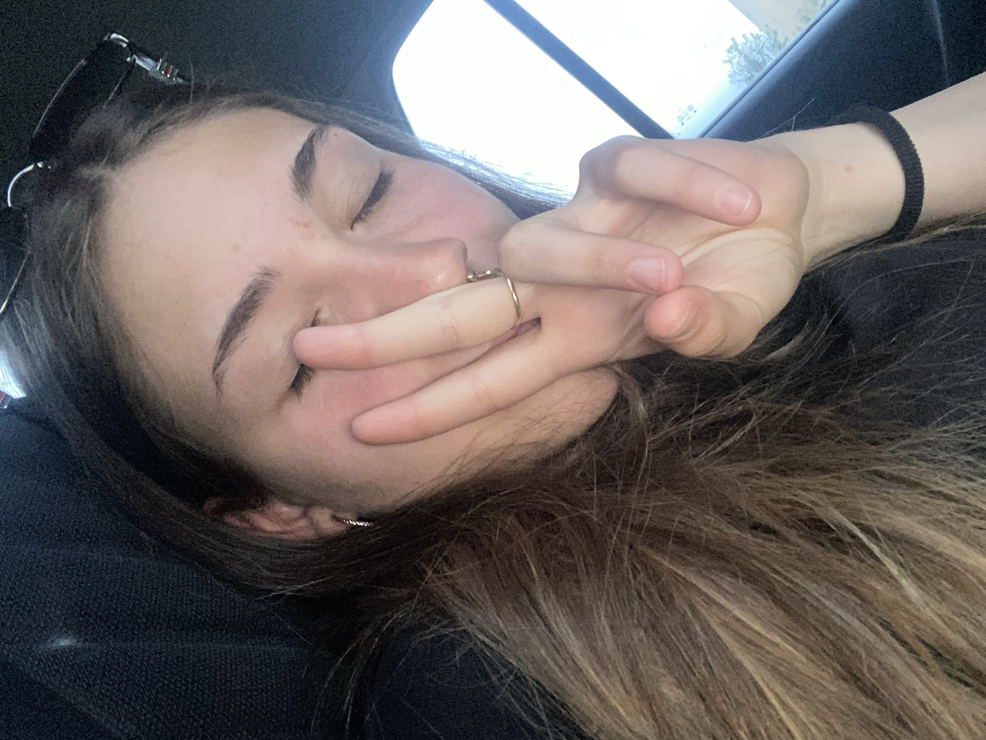 A close-up image shows a fair-skinned person, presumably female, with closed eyes and brown hair, possibly resting in a car. Her hand is partially covering her face, and a gold ring is visible on one of her fingers. Sunglasses are placed on her head near the top of the frame. A black wristband is on her wrist. The background shows a car window and hints of the outside environment. There is no identifiable location information suggesting Debelt, Bulgaria.