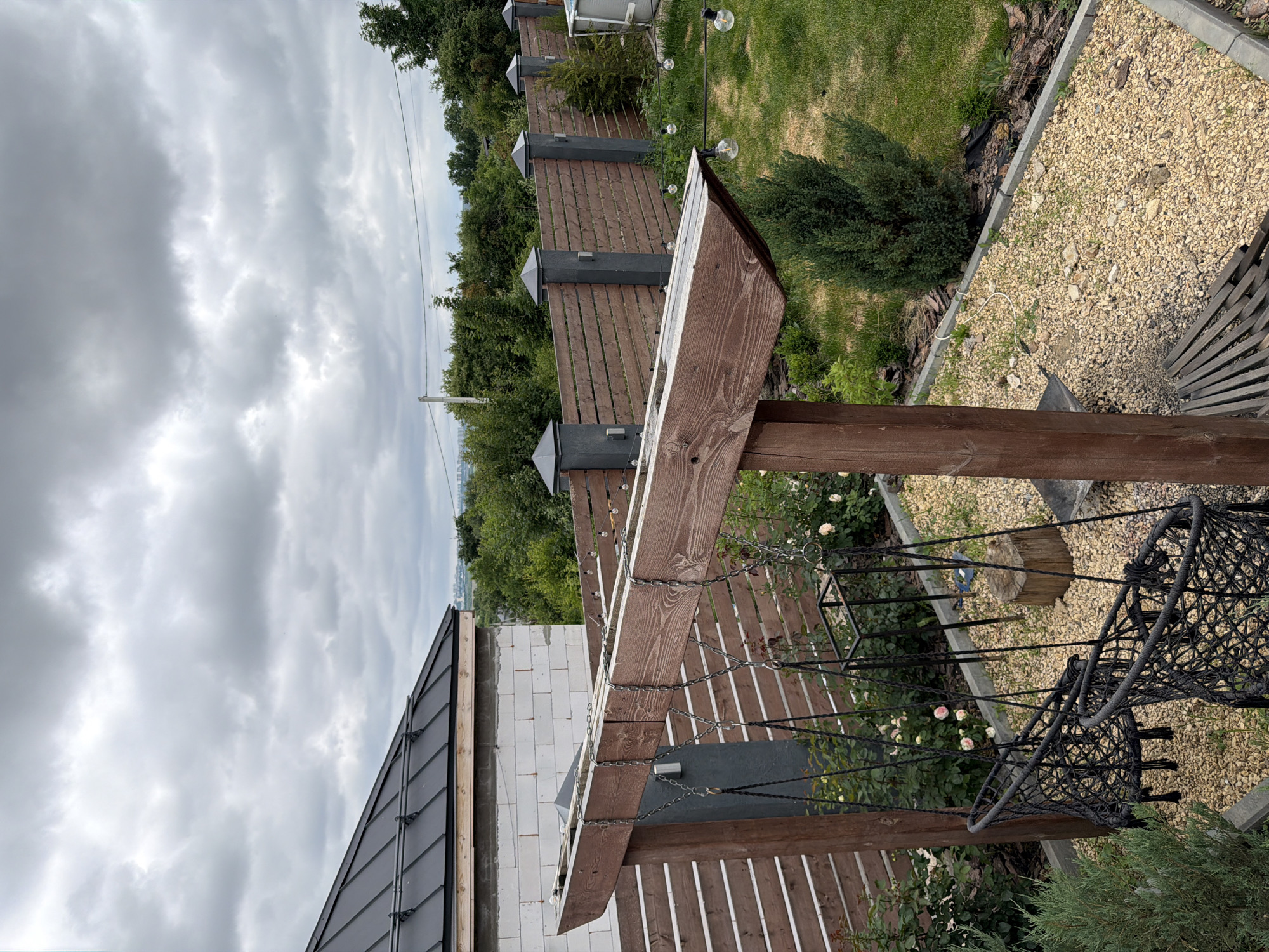 The image depicts a backyard scene in Semiluki, Russia. In the foreground, a wooden pergola supports two macrame swings hanging from metal chains. The swings are black and appear empty. To the right of the swings, a gravel area is visible, with some scattered stones and a wooden log. Beyond the pergola, a horizontal wooden fence with gray posts separates the yard from the surrounding area. The fence is adorned with string lights featuring clear bulbs. Behind the fence, lush green trees and bushes form a dense backdrop. A portion of a white brick house with a dark metal roof is visible in the upper left corner of the image. The sky is overcast with heavy, gray clouds. The overall scene suggests a residential outdoor space, possibly a garden or patio area.