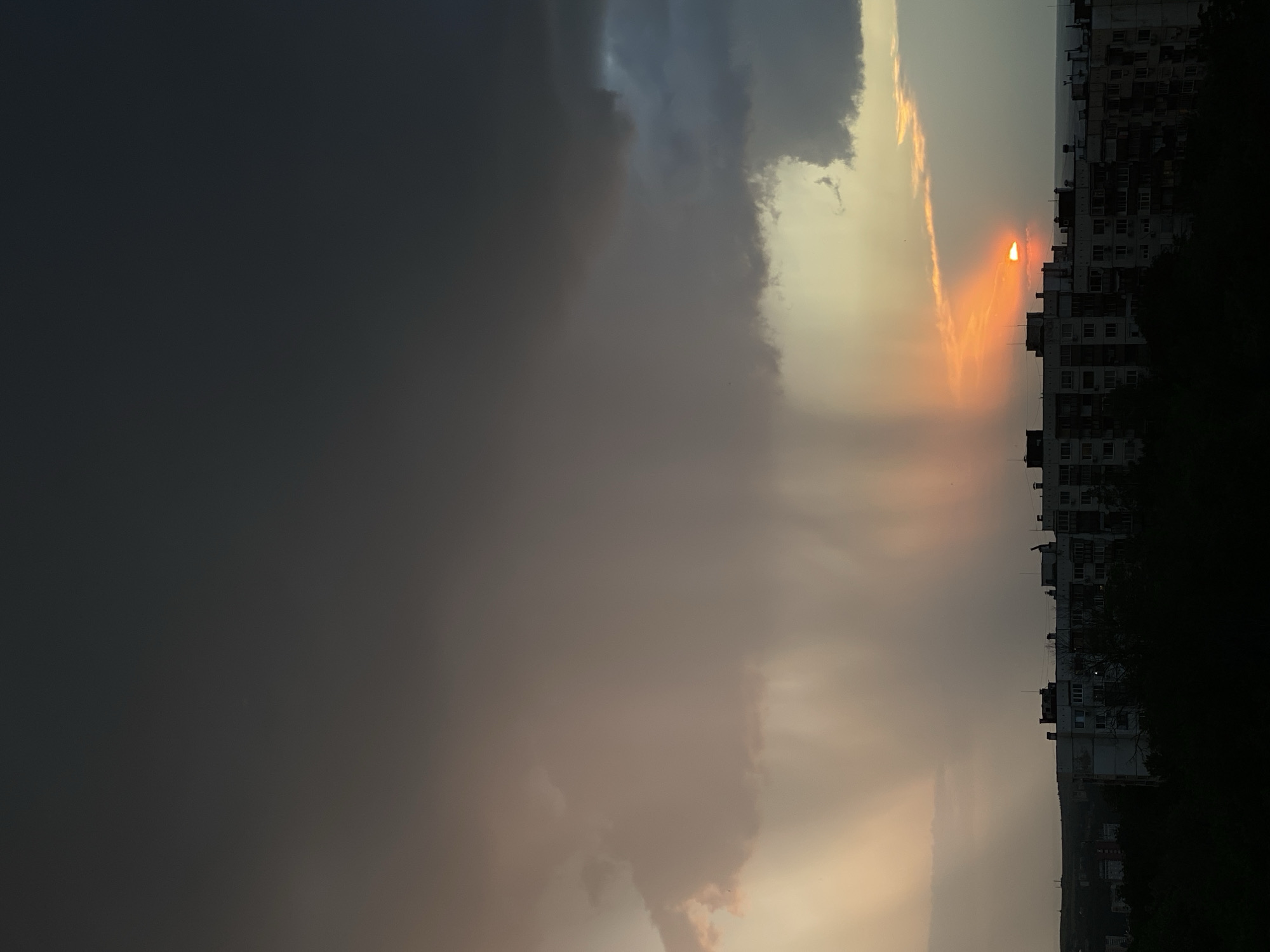 The image shows a sunset in the city of Pleven, Bulgaria. The sun is partially obscured by clouds, but its orange glow is visible near a tall building with many windows. Dark storm clouds fill the left side of the image. The silhouette of trees is visible along the bottom right edge of the image. There are no people visible in the image.
