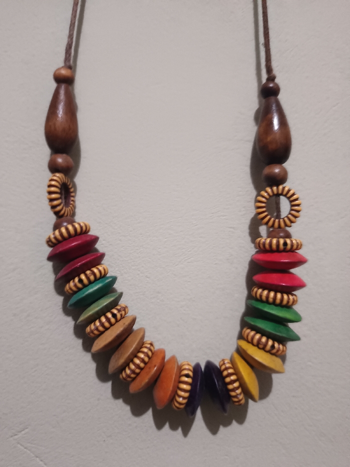 This image, captured in Gotha, Germany, displays a wooden bead necklace suspended against a plain, light grey wall. The necklace forms a symmetrical U-shape. The suspension mechanism is a dark brown braided cord, approximately 2mm in diameter. The cord connects to two identical, polished, dark brown wooden beads, each roughly 4cm long and teardrop-shaped, tapering towards the top. Below these, on each side, is a smaller, spherical dark brown bead, followed by a light tan, ribbed, ring-shaped wooden bead, roughly 1.5cm in diameter, exhibiting dark horizontal striations.
The main body of the necklace features a sequence of disc-shaped wooden beads, approximately 2.5cm in diameter and 0.5cm thick, interspaced with smaller, barrel-shaped light tan wooden beads, approximately 1cm long, also exhibiting dark horizontal striations. The disc-shaped beads are painted in various solid colors: red, dark green, olive green, light brown, orange, dark purple, and yellow, arranged in an alternating pattern along the necklace's lower arc. The light source appears to be from the upper left, casting distinct shadows of the necklace onto the wall to its right and below. No other objects, people, or environmental details are visible.