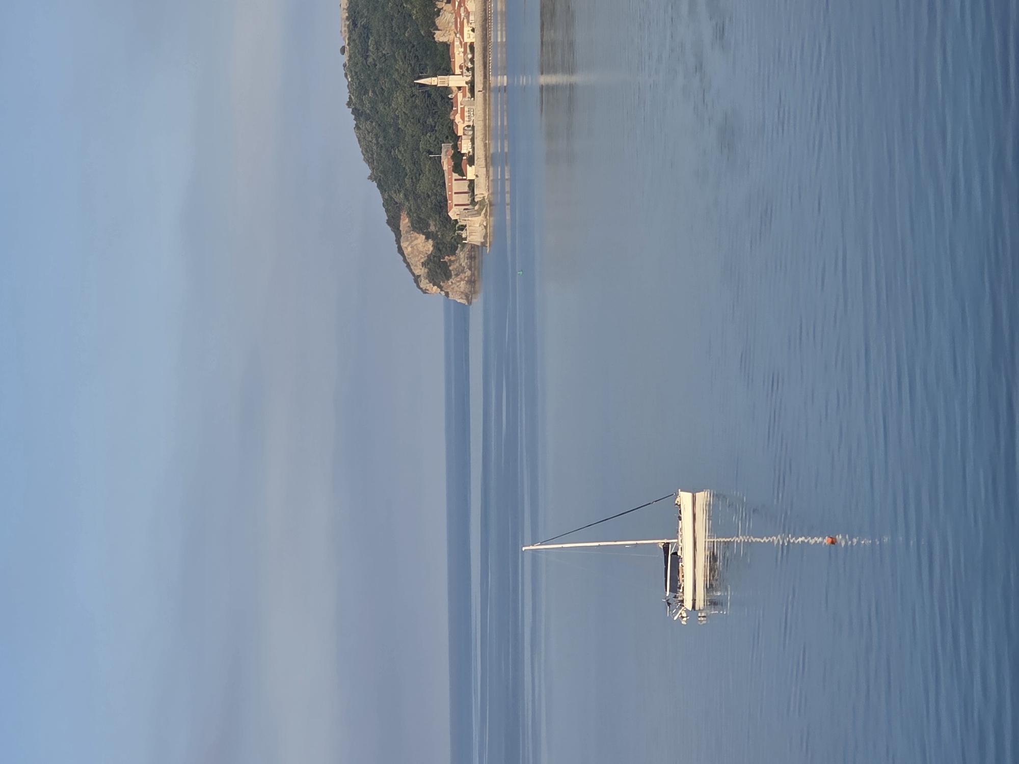 An eye-level shot captures a serene view of the Adriatic Sea with a sailboat and the historic old town of Budva, Montenegro in the background. The sky is a pale, hazy blue, with a faint cloud band across the horizon. The sea is calm, reflecting the sky and surrounding elements with gentle ripples.
In the foreground, a white sailboat with a single mast and a dark sail is anchored in the water, its reflection clearly visible below. A small orange buoy is attached to a rope extending from the boat.
To the right, the peninsula of Budva's old town rises from the sea. A lush, green forested hill forms the backdrop for a cluster of light-colored buildings with terracotta roofs, characteristic of Mediterranean architecture. A tall, slender church spire is prominently visible among the buildings. The stone walls of the old town are visible along the coastline. The overall impression is one of tranquility and natural beauty, highlighting the architectural heritage of Budva against the calm sea.