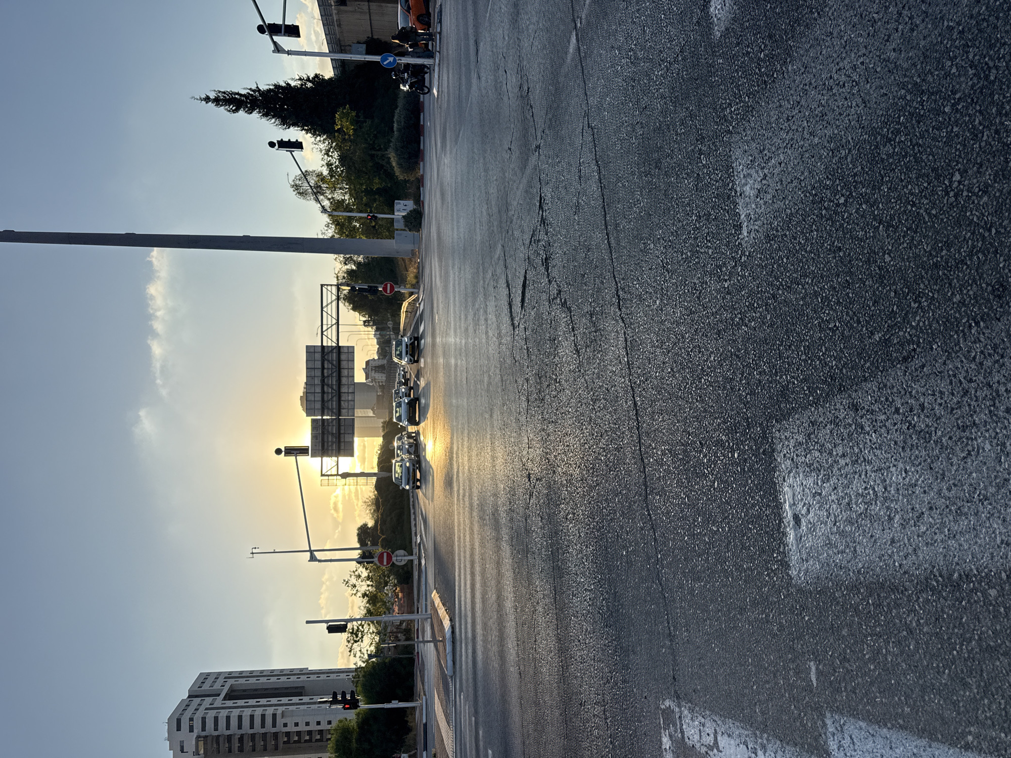This image depicts an intersection in Hod HaSharon, Israel, bathed in the warm glow of the setting sun. The foreground is dominated by a wet asphalt road with visible cracks and faded white pedestrian crossings. In the midground, several cars are stopped at a traffic light, their headlights reflecting on the wet surface. The background features a tall, modern residential building on the left, and a dense cluster of trees on the right, including a prominent dark green conifer. Traffic lights are visible at the intersection, with some showing red signals. A large overhead road sign structure spans across the intersection. The sky is a clear blue with a few wispy clouds, and the sun is low on the horizon, casting long shadows and illuminating the scene with a golden hue. There are no people clearly visible in the image, though a motorcycle with two occupants is present near the right side of the frame, along with a person standing next to an orange car.