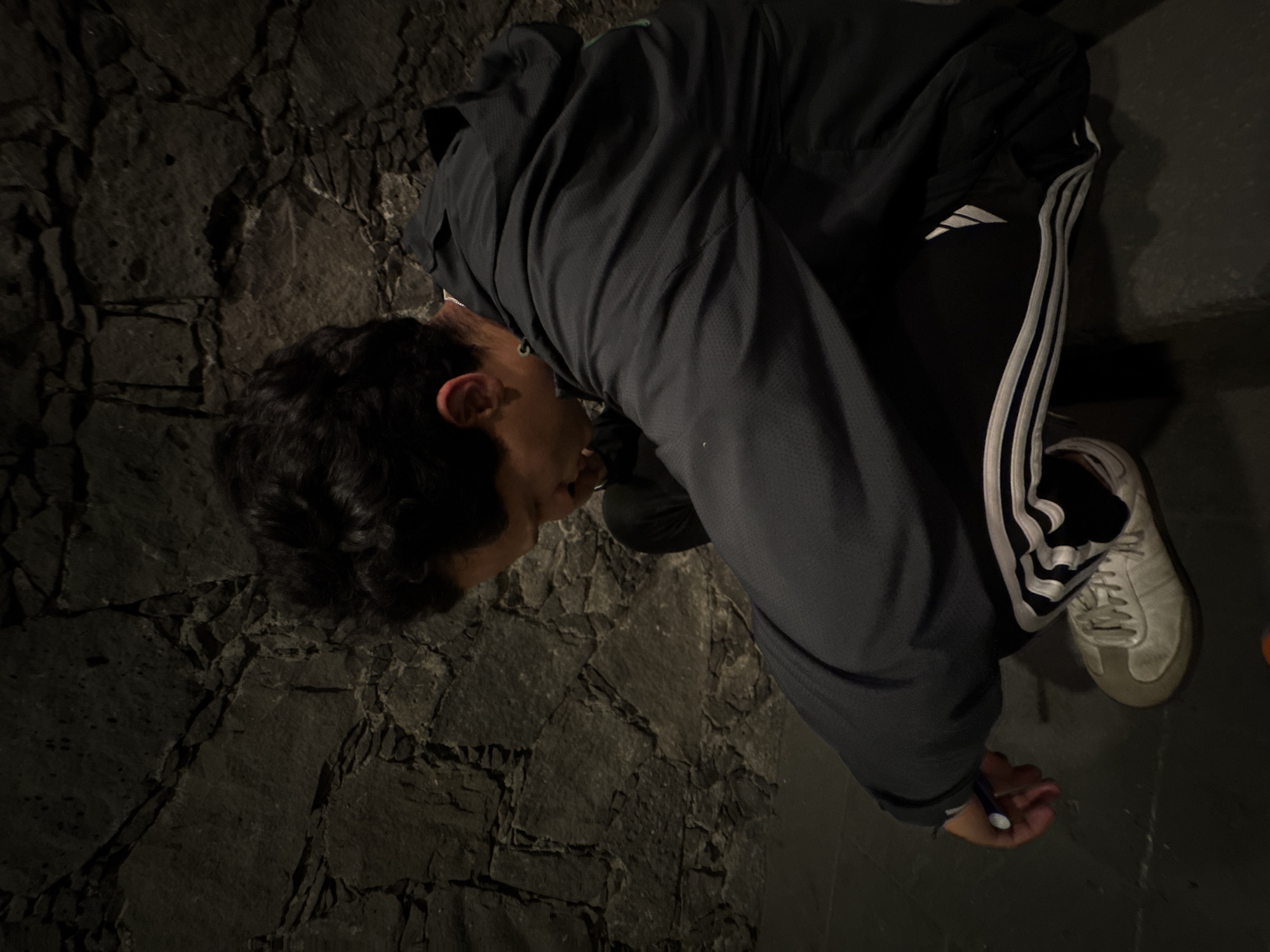 The image, taken in Mexico City, Mexico, features a person captured from a slightly elevated side angle, bent over and looking downwards. The individual is wearing a dark grey or black athletic jacket with a textured surface and matching dark pants adorned with the distinctive white triple stripes and an Adidas logo on the side. They are also wearing light-colored sneakers.
The setting is characterized by a rugged, dark stone surface, appearing as a wall or uneven ground composed of irregularly shaped, dark grey rocks, possibly of volcanic origin. The dim lighting suggests an indoor space, a subterranean area, or an outdoor scene at night. The person's posture indicates they are engaged in an activity requiring close attention to something below them. In their right hand, they hold a small object that appears to be a white and blue pen or marker, implying they may be writing, drawing, or making a mark on the unseen surface. The specific time of day or weather is not discernible, and no particular emotions are visible from this angle.