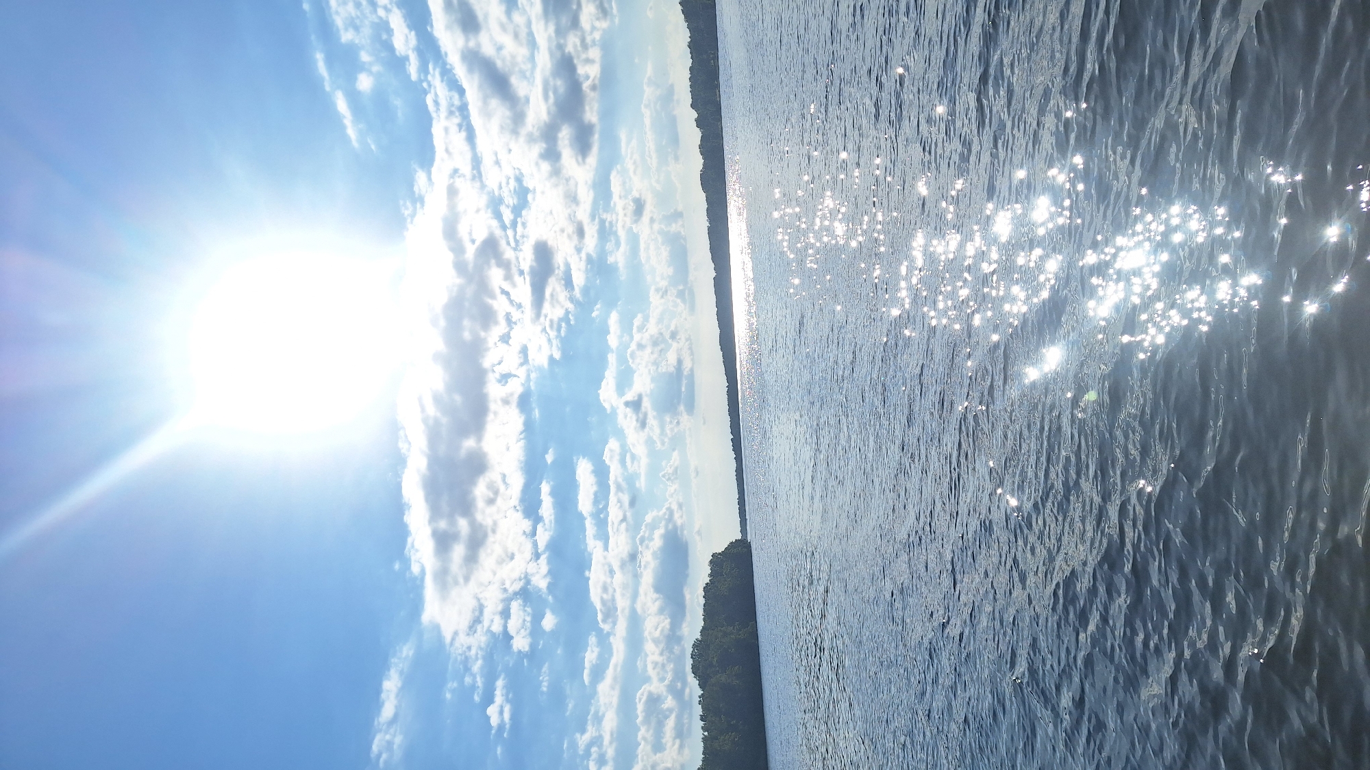 The image shows a sunlit body of water, likely a river or lake, with a distant tree-lined shore under a bright, partly cloudy sky. The sun is prominently positioned in the upper left quadrant, casting brilliant reflections across the rippling water surface. The scene depicts a natural outdoor environment, and the location is identified as the Daugava River in Koknese, Latvia. There are no people or specific man-made structures visible, emphasizing the serene and natural aspect of the landscape. The water is calm with gentle waves, and the sky is a clear blue interspersed with scattered white clouds. The overall impression is one of a bright, sunny day in a tranquil setting.