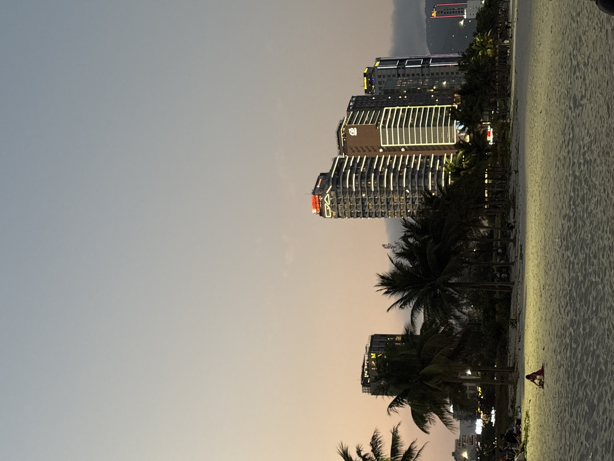The image captures a vibrant urban beach scene in Đà Nẵng, Vietnam, at dusk or early evening. The sky displays a subtle gradient, transitioning from a pale, warm orange-pink near the horizon to a cool, light grey-blue overhead, indicating the fading daylight.
In the foreground, a broad expanse of light-colored sand stretches across the bottom portion of the frame. Numerous dark silhouettes of palm trees line the edge of the beach, creating a tropical atmosphere. Several people are visible on the sand, including a distinct figure in bright red or pink attire sitting or kneeling towards the right side. Other smaller groups are indistinctly visible further down the beach, enjoying the evening.
The midground features an array of modern, multi-story buildings, likely hotels or resorts, illuminated with numerous bright white and yellow lights that outline their structures and balconies. One prominent building clearly displays the "Hilton Garden Inn" logo in red on its facade, along with its characteristic branding. Another tall building to the left also shows illuminated elements and a logo.
In the far distance, subtle outlines of mountains are visible against the darkening sky, with a distinct red-lit structure, possibly a tower or part of a building, standing out on a distant peak or high-rise. The scene conveys a peaceful yet urban coastal setting, characteristic of a lively city at twilight.