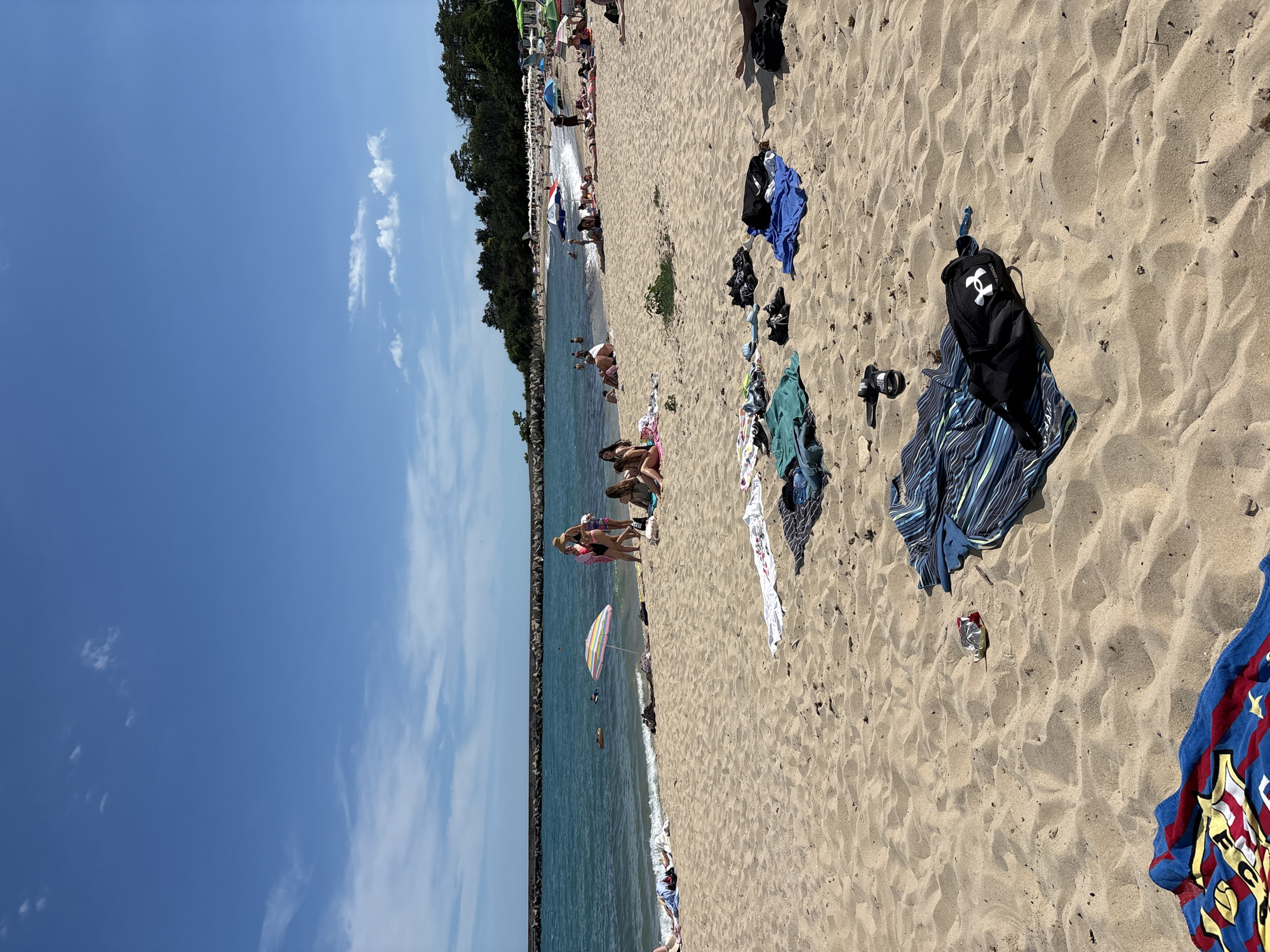 This is a daytime, eye-level, full shot of a crowded sandy beach in Varna, Bulgaria, with the sea in the background. The water is a deep blue with small waves crashing on the shore. A long, dark stone breakwater stretches across the horizon.
On the beach, there are numerous people sunbathing and swimming. Several towels and bags are scattered on the sand, indicating people are spread out across the beach. Some individuals are gathered near the water's edge, while others are further up on the sand. A colorful umbrella provides shade for a small group. In the foreground, a variety of beach towels and personal items, including a black backpack with an "H" logo, are visible on the sand. The sky is a clear blue with a few scattered white clouds.