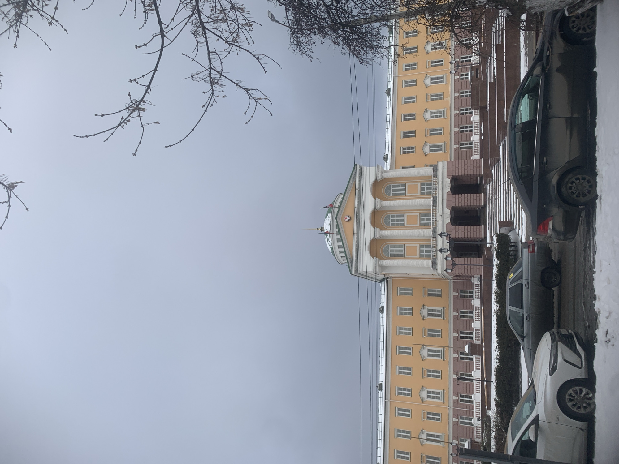 The image shows a street scene in Izhevsk, Russia, featuring a prominent yellow and white building with a dome. The building has multiple floors with numerous windows and a symmetrical facade. A red flag is visible atop the dome. In the foreground, several parked cars line the street, which appears to be covered in snow. Bare tree branches extend into the frame on the left side, contrasting with the overcast sky. Power lines run along the side of the building. The overall impression is a typical urban scene in a Russian city during the colder months.