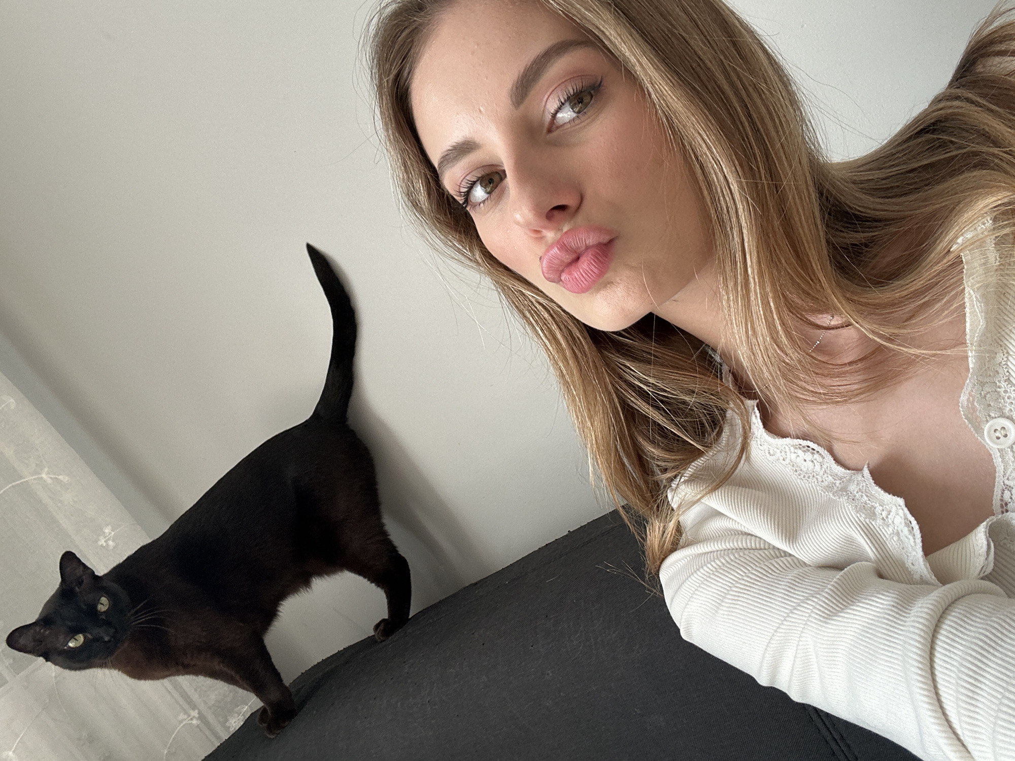 A woman poses for a selfie indoors with a dark brown cat. The woman has long, light brown hair and is wearing a white, long-sleeved shirt with lace trim. She is looking at the camera with her lips puckered. The cat is standing on a dark-colored surface, possibly a couch, and is looking at the camera. The background is a plain white wall. There is no indication this picture was taken in Warsaw, Poland.