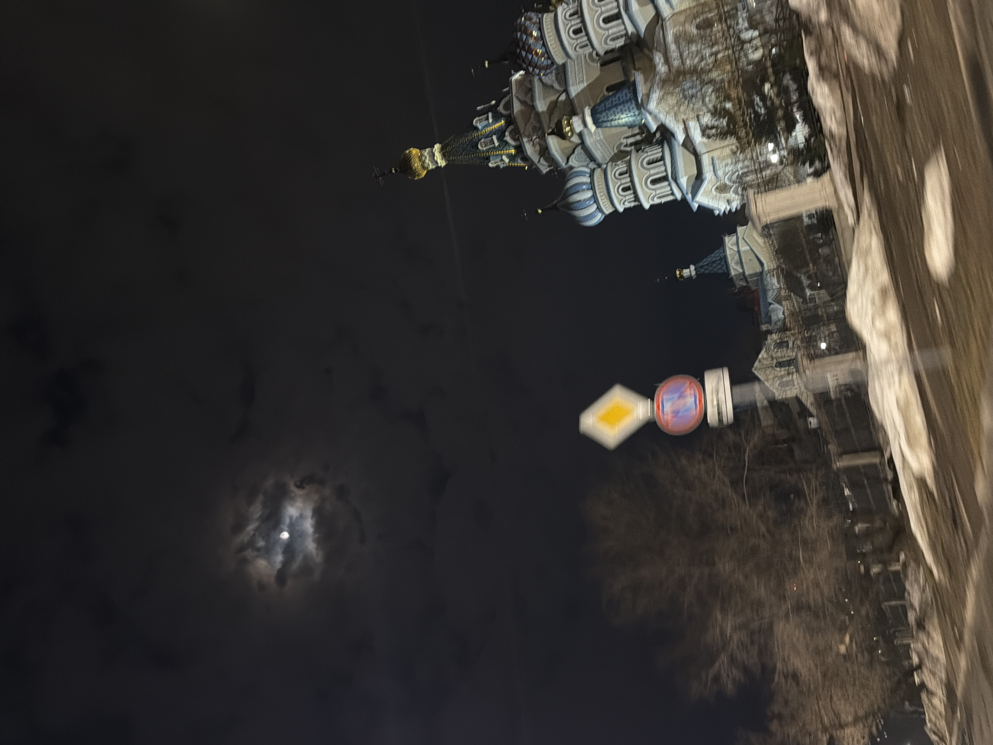 The image shows a night scene in Izhevsk, Russia. In the right half of the image is the brightly lit Mikhailovsky Cathedral. The background to the left shows a dark, cloudy sky with a visible moon. In the mid-ground is a traffic sign with a "No Entry" prohibition. The ground is not visible. The image is rotated 90 degrees counter-clockwise.