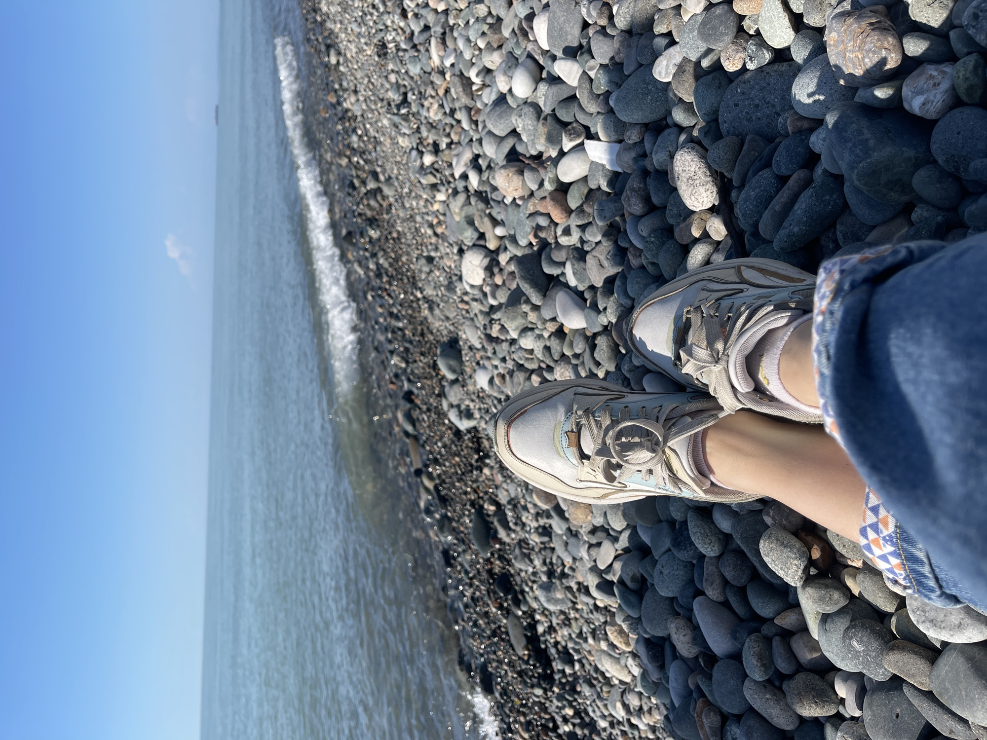 This image, possibly captured in Batumi, Georgia, presents an individual standing on a pebble beach. The foreground consists of a collection of predominantly gray and dark gray, smooth, rounded pebbles of various sizes, scattered along the shoreline. Overlapping the stones are the feet of a person wearing beige athletic shoes and socks. The person is wearing blue jeans. The middle ground shows the waterline with gentle waves lapping at the shore. Beyond the waterline, the view extends to the horizon where the sky transitions from blue to a paler tone near the horizon. The sky is cloudless, indicative of clear weather. The overall composition suggests a moment captured at a beach near Batumi.