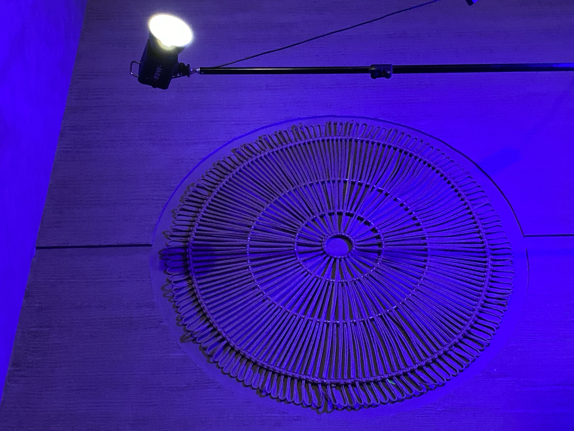 A round, wicker placemat sits under artificial lighting in what appears to be a room, likely indoors. The light source, an 80W lamp on an adjustable arm, is positioned above and to the left, casting a bright, focused light. The entire scene is bathed in an artificial blue light, which could be from the lamp or another source. The surface beneath the placemat appears to be a wooden panel. There is no indication in the image as to whether this is Izhevsk, Russia.