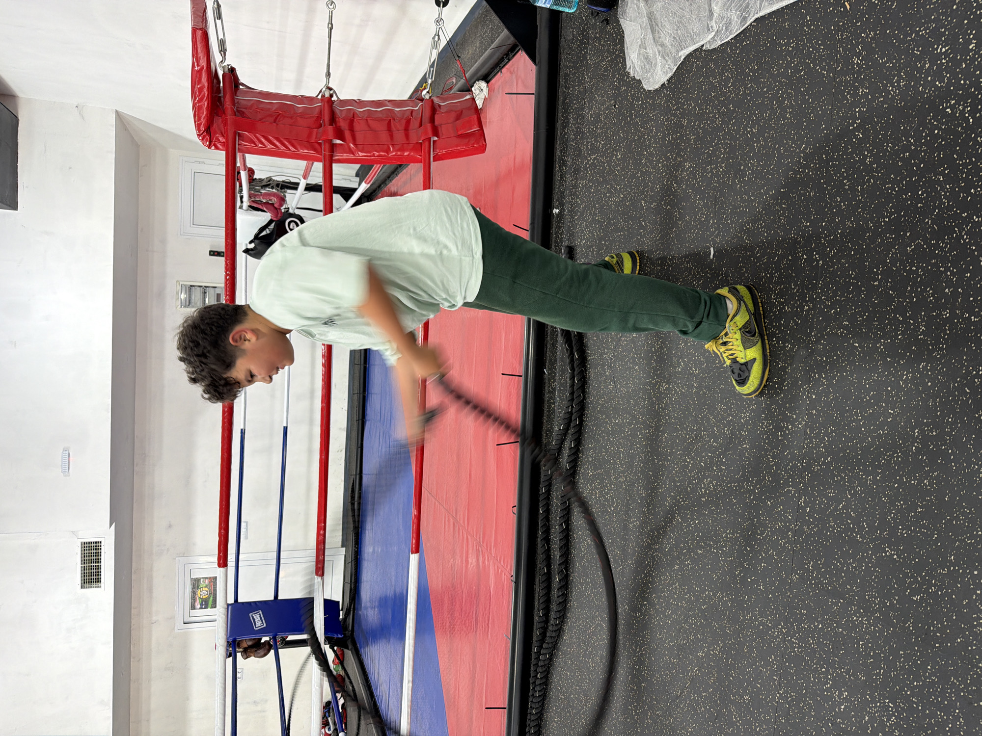 A boy is using battle ropes in a gym. The gym has a boxing ring in the background. The boy is wearing a light green t-shirt, dark green pants, and bright green and black shoes. The gym has a black floor with white speckles. The location is not evident from this image.