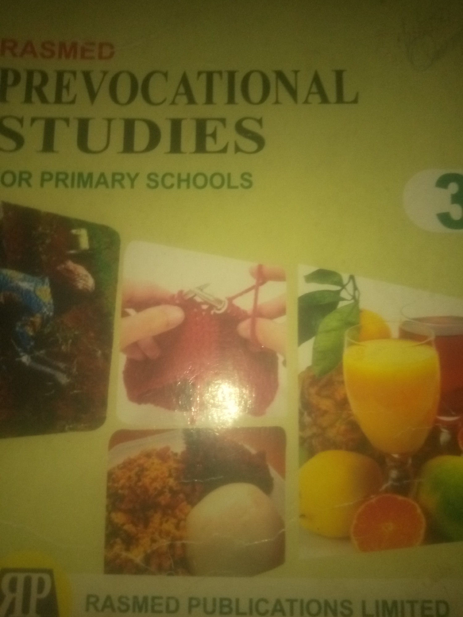The image is a close-up, eye-level, medium shot of the cover of a book titled "RASMED PREVOCATIONAL STUDIES FOR PRIMARY SCHOOLS". The background is a pale yellow-green. The title is printed in large, dark green capital letters. "RASMED" is at the top, followed by "PREVOCATIONAL", and then "STUDIES". Below this, in smaller capital letters, is "FOR PRIMARY SCHOOLS". To the right of the title, a large green "3" is enclosed in a light gray oval.
The lower half of the cover features a collage of four rectangular images. The top left image shows a dark brown, slightly blurred scene with a person's hand visible, and possibly some soil or grass. The top right image depicts hands knitting with red yarn. The bottom left image contains a close-up of food, appearing to be rice and a pale oval-shaped item. The bottom right image displays various fruits, including what looks like an orange or grapefruit slice, lemons, and a glass of orange juice. A logo with the letters "RP" in a stylized font is in the bottom left corner of the cover, above the text "RASMED PUBLICATIONS LIMITED" which is printed in dark gray capital letters across the bottom of the cover. The lighting appears to be artificial and slightly warm, creating some glare and reflections on the cover.