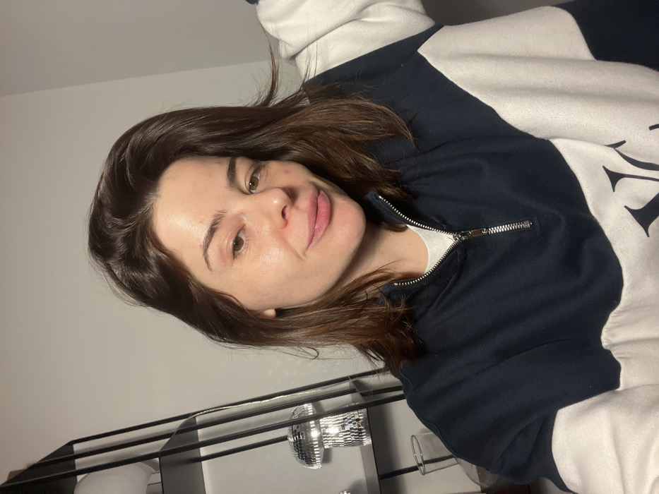 A woman poses indoors wearing a navy blue and white pullover with a partial zipper. Her shoulder-length brown hair is parted to the side. Her face is smooth and without makeup. A black metal shelf is visible below, featuring a disco ball and a white spherical object. The background is a plain, light-colored wall. The image is slightly tilted to the left.