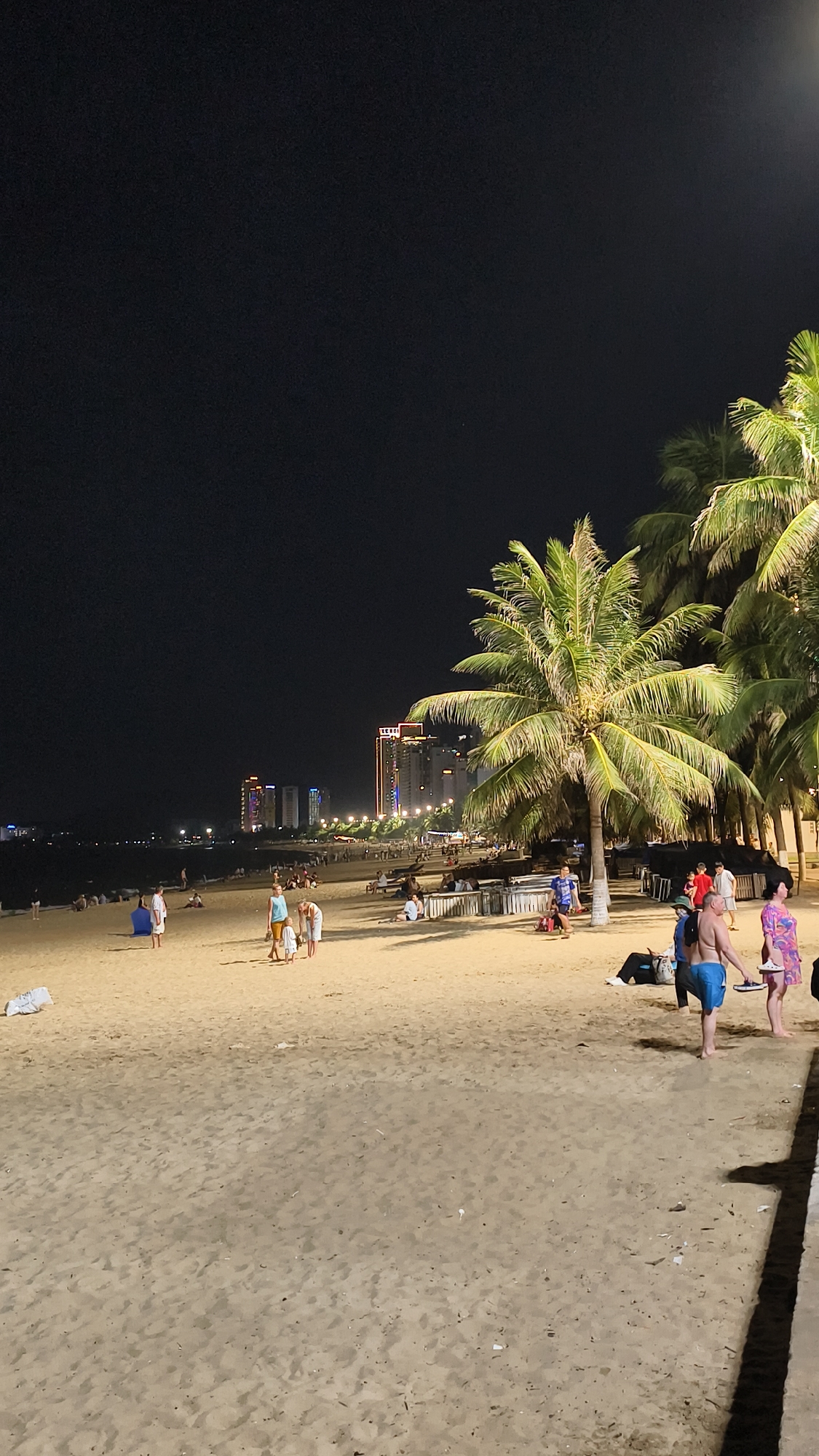 An aerial view of a sandy beach at night showcases a lively scene with scattered palm trees, distant city lights, and numerous people enjoying the evening. The sand is a light tan, with footprints and scattered debris visible. In the foreground, a group of people are walking and standing on the beach, some wearing swimwear. Further back, more individuals are seated or walking, enjoying the nighttime ambiance. The background is dominated by a dark, starless sky, punctuated by the glowing lights of a city skyline featuring tall buildings illuminated with colorful lights. The presence of palm trees, some with their fronds lit by artificial light, adds a tropical feel to the urban coastal setting.