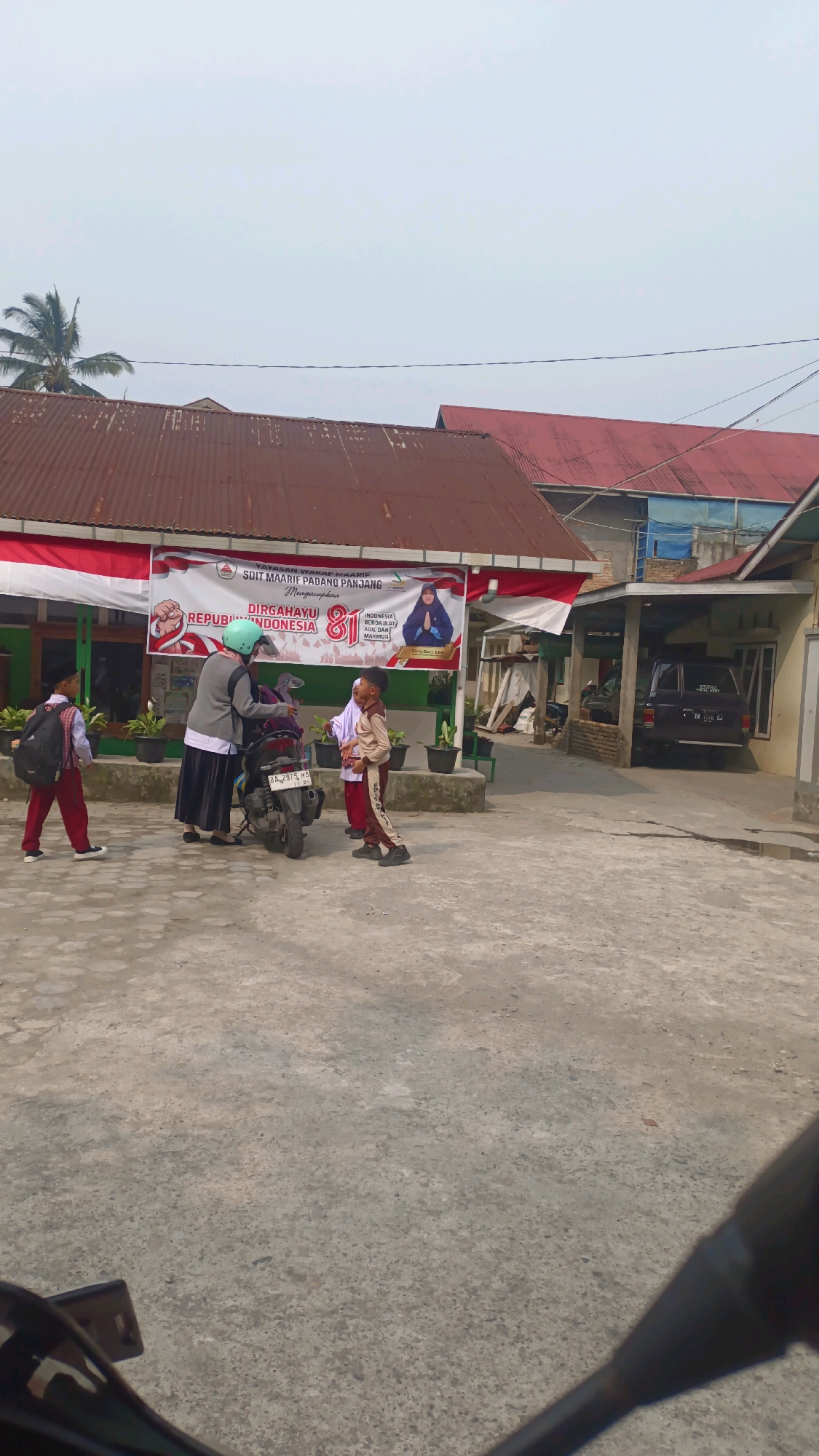 A group of people, including at least two children in school uniforms and an adult woman wearing a hijab and a helmet, are gathered near a motorcycle in front of a building with a large banner. The banner, draped across the front of the building, displays Indonesian national colors (red and white) and text that appears to be a congratulatory message, possibly for an anniversary or independence day, given the presence of "DIRGAHAYU REPUBLIK INDONESIA 81". The building has a rusty corrugated metal roof and appears to be a school or educational institution. A dark-colored SUV is parked under a covered area to the right of the main building. The ground is paved with concrete. The scene suggests a moment of departure or arrival at the school, possibly in the morning or afternoon. The presence of the banner and the Indonesian flag colors indicate a festive or patriotic occasion. The overall environment is an outdoor, somewhat overcast, urban or suburban setting in Padang Panjang, Indonesia.