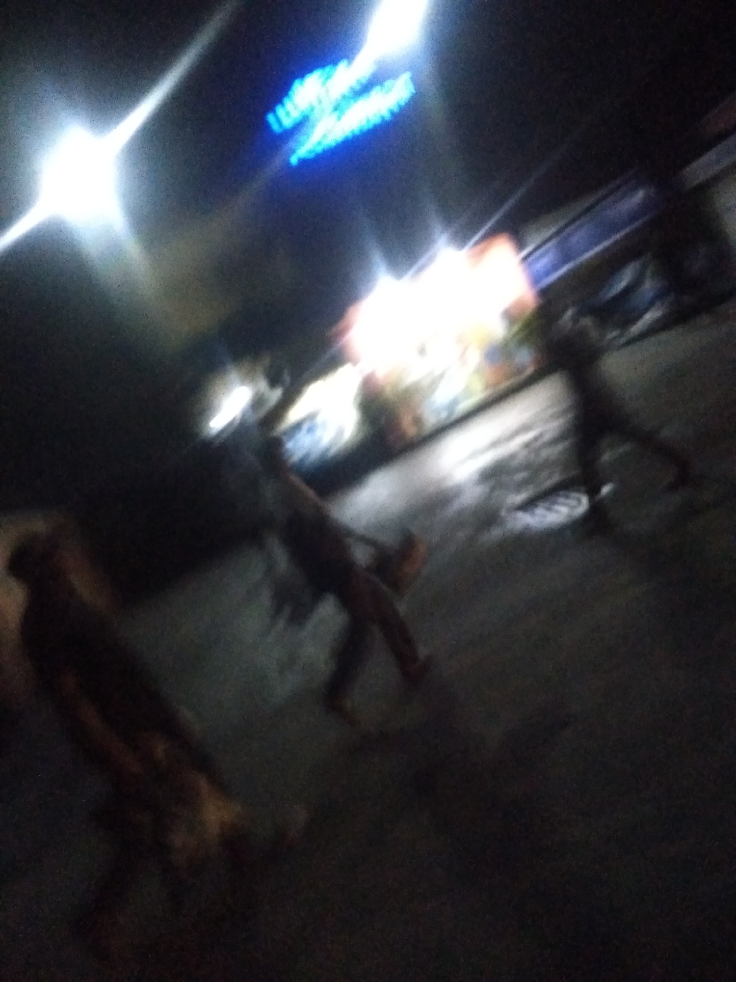 This very blurry and dark photograph captures a nighttime scene, likely in Shangisha, Nigeria, depicting an outdoor street or market area. Several human figures are present, heavily blurred by motion or low light conditions, rendering them indistinct silhouettes.
On the far left, a bright, overexposed light source dominates the frame, possibly a streetlight or vehicle headlamp. In the upper center, a highly blurred blue neon sign is visible, its glow hinting at text or a logo, though no specific words can be discerned. Below this, diffused, warmer lights emanate from what appears to be a shopfront or stall, displaying indistinct shapes and colors.
Three main human figures can be identified. One on the far left is a dark, elongated silhouette. Another figure in the middle is walking, with legs and torso partially visible, and appears to be carrying something low on their side. A third, equally blurred figure is seen on the right, primarily as indistinct legs. The ground beneath them appears to be a paved surface, reflecting some of the overhead light, which might indicate dampness. The image conveys a sense of movement and activity in a dimly lit, possibly bustling, urban or semi-urban environment at night.