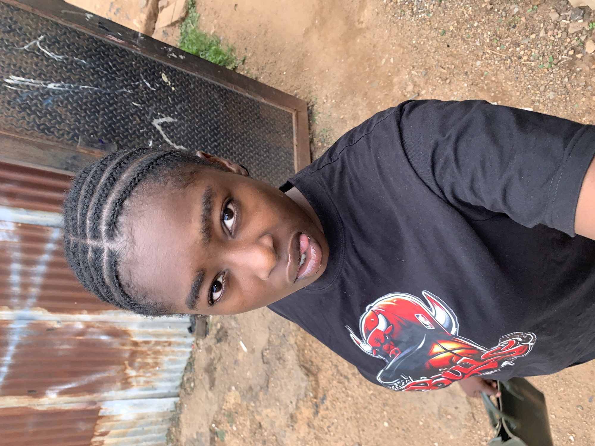 A young woman with braided hair, wearing a black t-shirt with a Chicago Bulls logo, is standing outside. The background consists of a corrugated metal wall, a weathered metal door, and dirt. The location is Rafin Par, Nigeria.