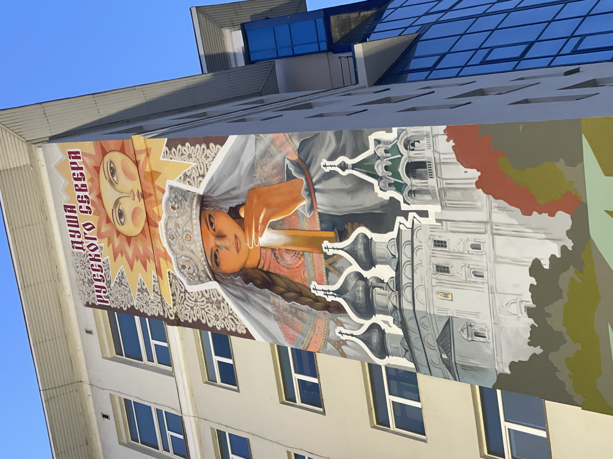A mural is painted on the side of a building. The mural depicts a young woman wearing a crown and a veil, holding a candle. Behind her, a sun with a face shines, and at the bottom, a church with multiple domes is shown. The text above the sun reads "ДУША РУССКОГО СЕВЕРА," which translates to "The Soul of the Russian North." This mural is located in Vologda, Russia, on the administrative building of the Vologda State Dairy Farming Academy, named after N.V. Vereshchagin.