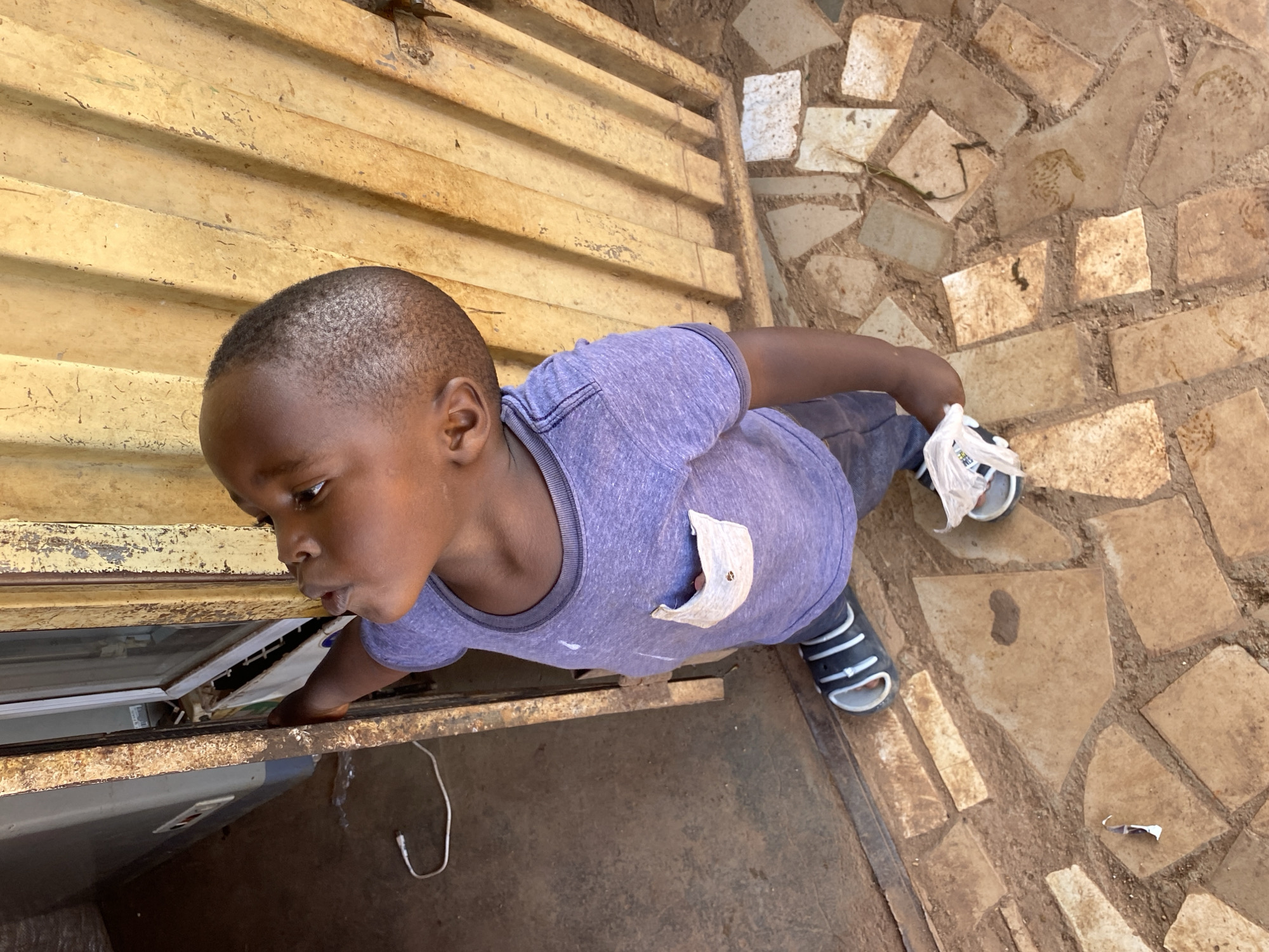 This overhead shot captures a young child, appearing to be male, leaning forward with an expression of focused curiosity. He has short dark hair and is wearing a purple short-sleeved t-shirt with a light-colored, slightly worn chest pocket, and dark trousers or shorts. On his feet are black and white striped sandals; the right sandal has a white plastic bag partially wrapped around it.
The child is intently looking down into an open cavity or section of what appears to be an appliance or a storage unit, revealing some internal components, wires, and metallic parts within. His left hand is resting on the lower edge of this opening.
To his left and above, there is a corrugated metal wall or surface, painted in a faded, yellowish-brown hue with visible rust and peeling paint. To his right and below, the ground is paved with irregular, earthy-toned tiles or flagstones, interspersed with dirt or cement. A white cable, possibly a charging cord, lies coiled on the dark ground directly below the opening the child is observing.
The lighting suggests it is daytime, casting bright, even illumination on the scene. The overall setting appears somewhat rustic or functional, potentially in a workshop or an outdoor/semi-outdoor area in Bukuru, Nigeria. No discernible text is visible in the image.