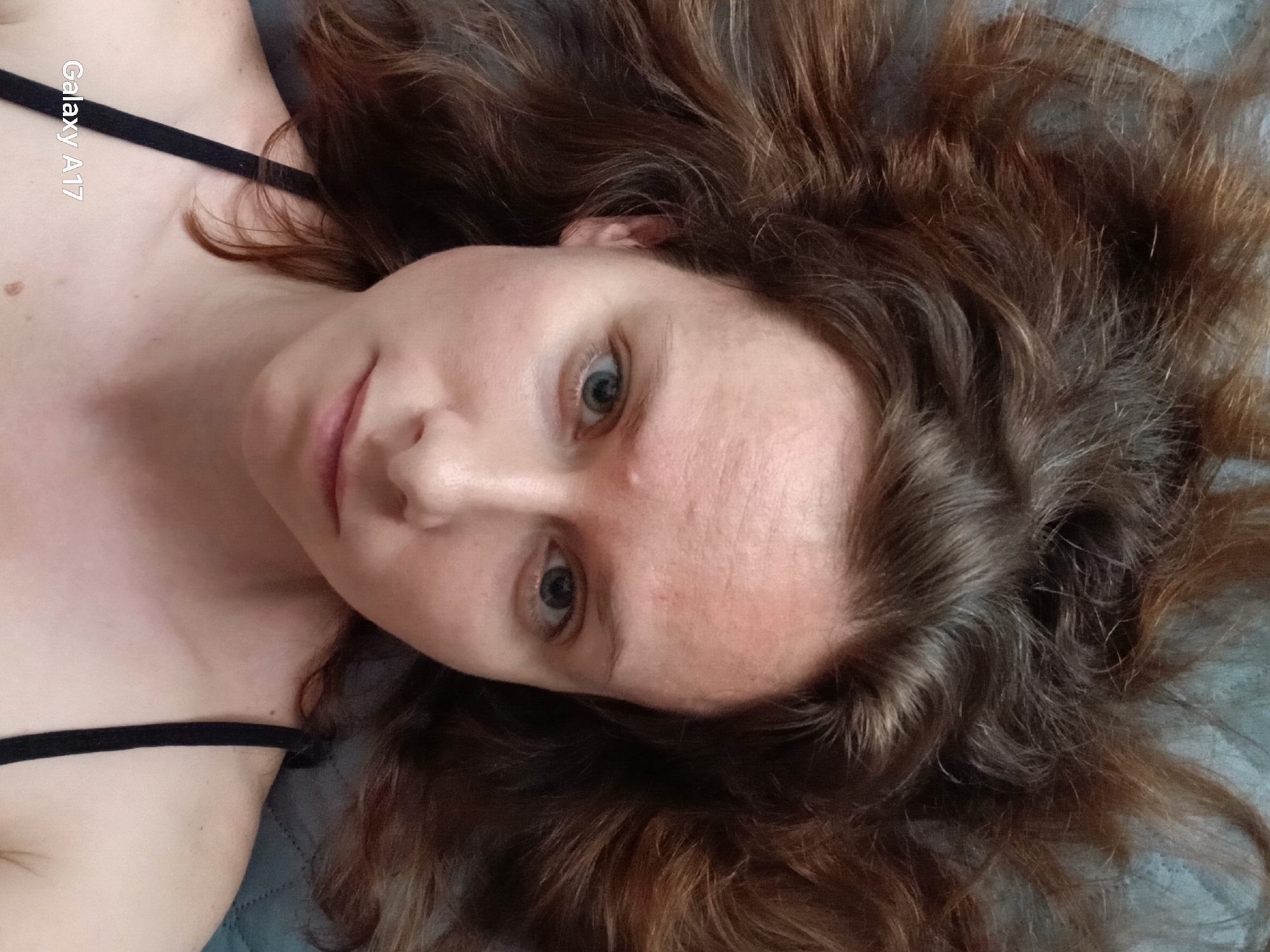 A woman with light skin and brown hair lies on a gray surface. She is facing upward with a slight smile. She is wearing a black strap on her shoulder. "Galaxy A17" is printed in white text on the left side of the image.
There is no indication of location in Yekaterinburg, Russia.