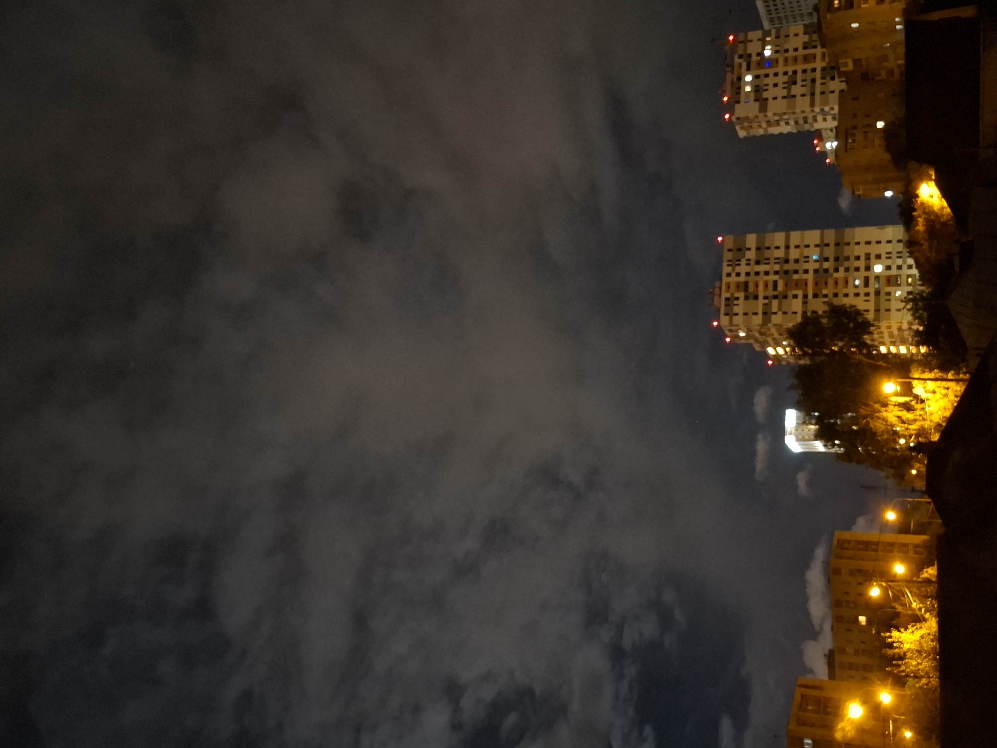 A nighttime cityscape scene in Tyumen, Russia, featuring several multi-story buildings and streetlights against a cloudy sky. In the foreground, the roofs of lower buildings are visible. Behind them, illuminated by streetlights, are trees and smaller residential buildings, some with lights on in windows. Further back, two prominent apartment buildings with distinctive checkered facades and red lights on their roofs stand out. To the right of these, another tall building is visible, brightly lit from within. The sky is dark with scattered clouds, some illuminated by the ambient city lights.