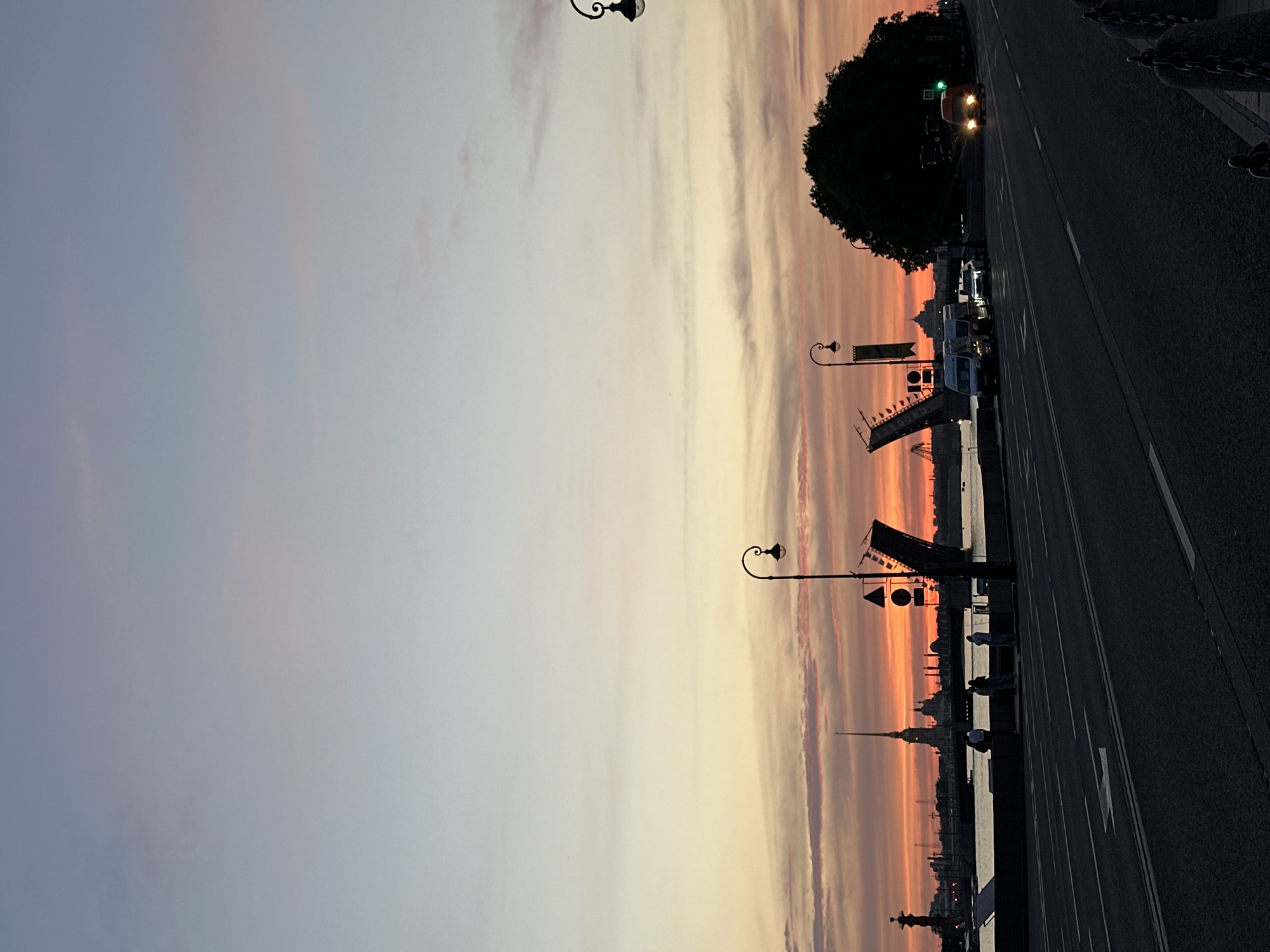The image depicts a sunset scene in an urban environment, likely a city with a prominent river or body of water. The sky is a gradient of colors, transitioning from pale blue and pink at the top to vibrant orange and red near the horizon. Silhouetted against the colorful sky are several landmarks, including spires and buildings, suggesting a historical or architectural significance. A bridge, partially raised, is visible in the mid-ground, indicating it is a drawbridge that can be opened for maritime traffic.
Along the waterfront, a few individuals are standing, appearing to observe the sunset or the city skyline. The foreground features a road with visible lane markings and a few vehicles. One vehicle, an orange van, has its headlights on and is approaching the camera. The scene is illuminated by the fading daylight and artificial lights from streetlamps and the vehicles. The overall atmosphere is tranquil and evocative of an evening cityscape. The specific location is unknown.