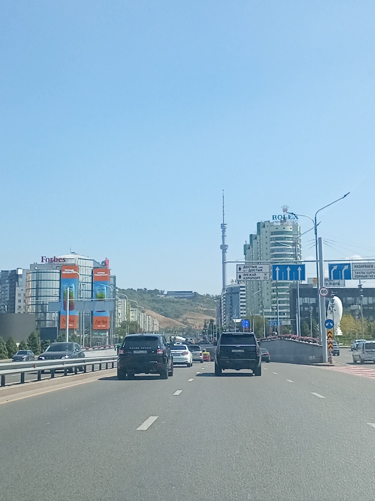The image shows a view from a vehicle on a multi-lane highway in Almaty, Kazakhstan. The highway is paved, with clear lane markings. Several cars are visible on the road, including black SUVs and a white car. On the left side of the highway, there is a concrete barrier and some trees. In the background, there are tall modern buildings. One building on the left has a sign that says "Forbes". Another prominent building in the center has a "ROLEX" sign on top and a tall tower behind it. There are also road signs indicating directions to "Достык" and "Аэропорт". The sky is clear and blue. The scene appears to be a busy urban environment.
