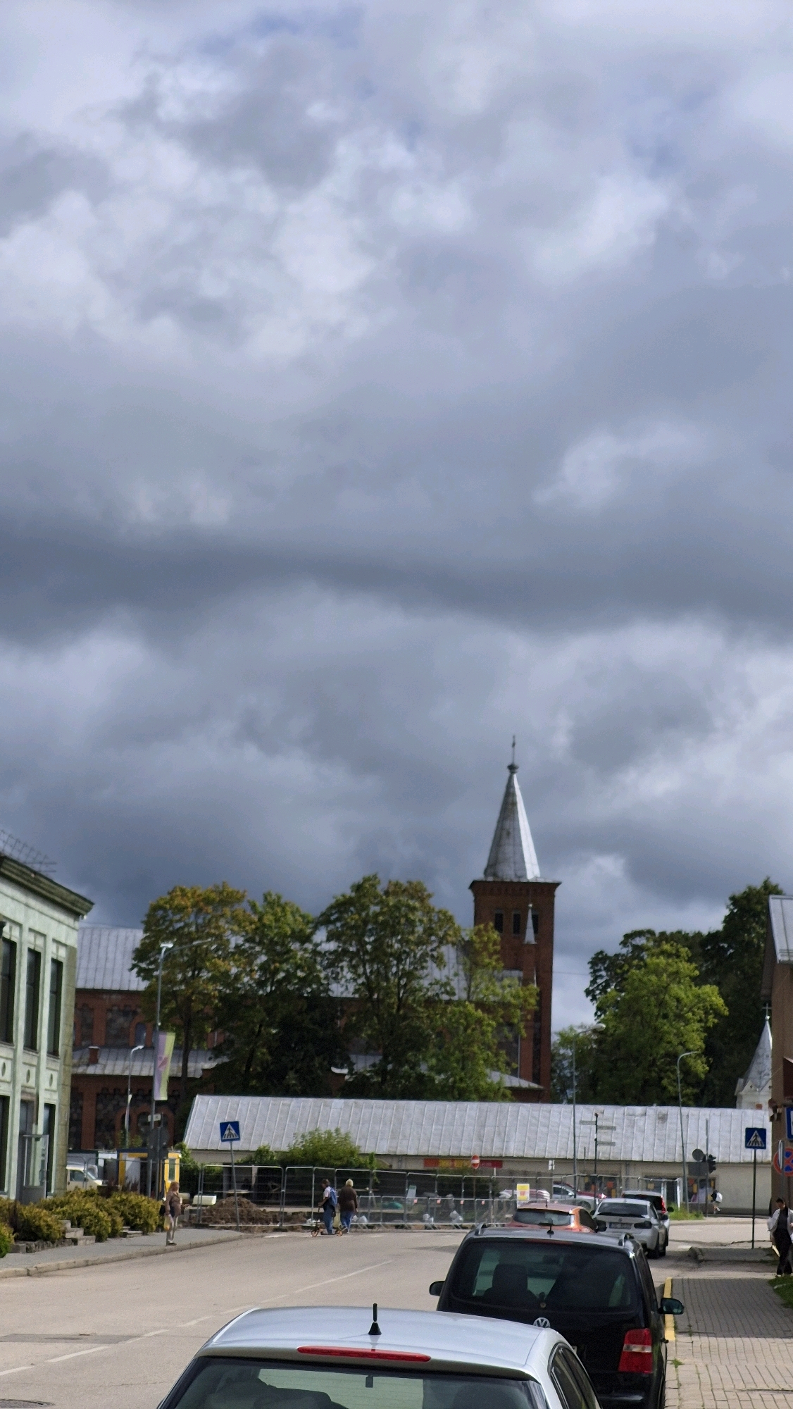 The image shows a street scene in Preiļi, Latvia, under a cloudy sky. In the background, a red brick church tower with a white spire stands out, flanked by trees. To the left, a light-colored building with multiple windows is visible. The street is lined with parked cars, and several people are walking or standing on the sidewalk. Construction barriers are set up across the street in the mid-ground, indicating ongoing work. The overall atmosphere is overcast, suggesting possibly inclement weather.
