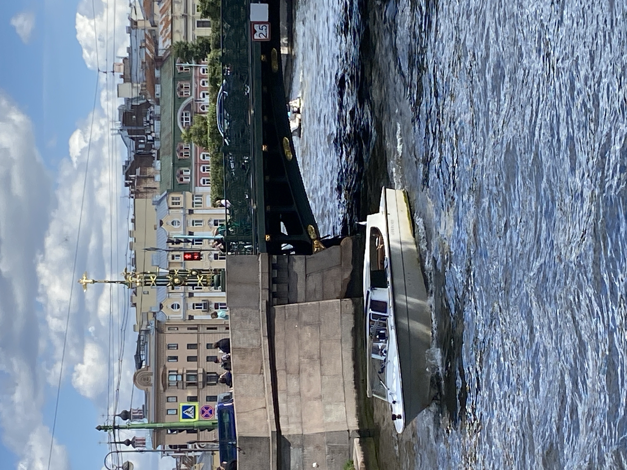 A white pleasure boat navigates a blue waterway under a green bridge in Saint Petersburg, Russia. The boat is positioned in the foreground of the image, moving from left to right, with its bow pointing towards the right. Its occupants are visible through the windows.
The bridge spans the waterway, with its underside casting a shadow on the water. Vehicles are visible on the bridge, including a dark sedan. Beyond the bridge, buildings with a mix of architectural styles, painted in various colors including beige, yellow, and green, line the city's waterfront. The sky above is blue with scattered white clouds.
On the left side of the image, a stone bridge support is visible, with people gathered on top, overlooking the water. A street light and traffic signals are also present near the bridge. The overall scene depicts a typical urban waterway environment in Saint Petersburg on a bright, cloudy day.