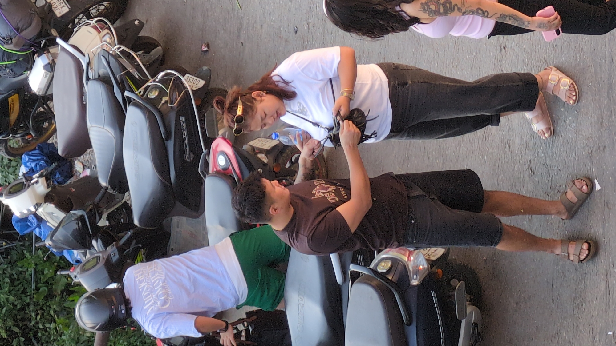 The image captures a scene in Bhagsu, India. Three people are visible amidst a parking area filled with motorbikes. A woman with sunglasses on her head, wearing a white t-shirt and dark jeans, is looking at a camera held by a man in a brown t-shirt and dark shorts, who is glancing up at her. A water bottle is between them. A third person, wearing a white long-sleeved shirt, green shorts, and a helmet, is partially visible next to the man. Another woman is partially seen on the upper right, wearing a pink top and dark pants, with a dragon tattoo on her left arm. The ground is paved.