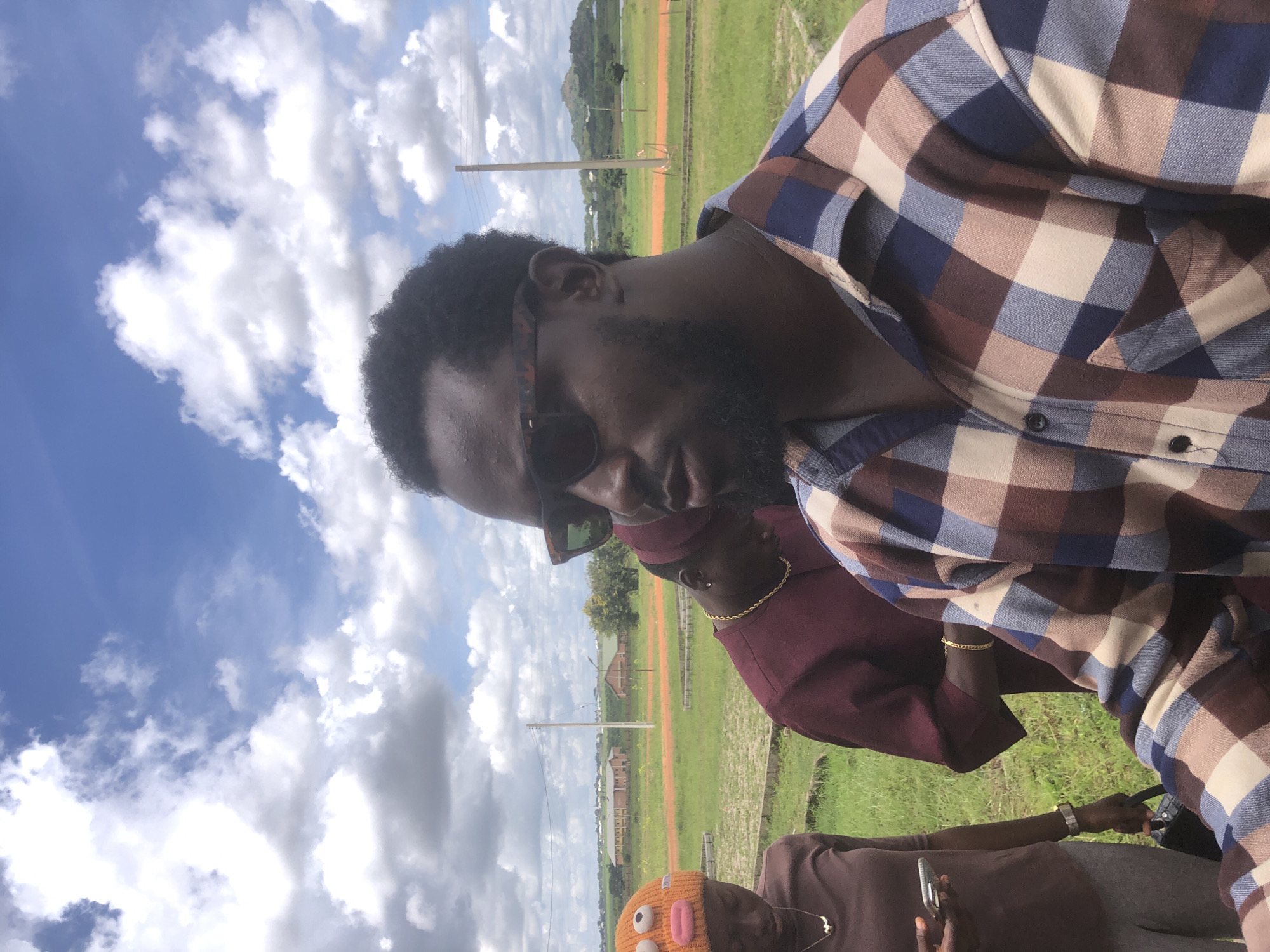 In Farin Yashe, Nigeria, under a cloudy sky, three individuals are visible outdoors in a grassy area with some construction in the background. The person in the foreground, a man with dark skin and a beard, is wearing sunglasses and a plaid shirt in shades of brown, blue, and tan. To his left, a person wearing a maroon shirt and a gold chain looks down. Further to the left, a person with dark skin wears an orange beanie with a cartoon face, a brown long-sleeved shirt, and grey pants. This individual holds a smartphone in their left hand and a black bag in their right. The background features a red dirt path, green grass, trees, utility poles, and buildings that appear to be part of a school or administrative complex. Hills are visible in the distance.