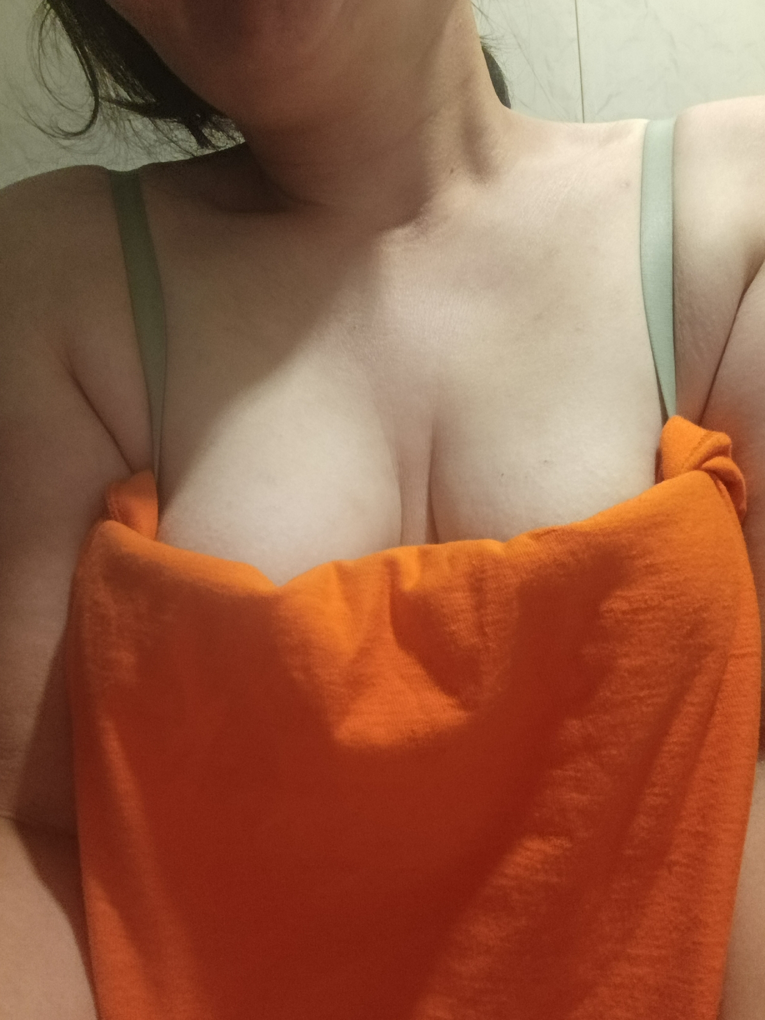 A torso shot of a woman with fair skin and dark hair. She's wearing a light green bra and an orange towel is wrapped around her chest, covering part of her breasts. The background appears to be a light-colored tile wall. There is no indication of location in the city of Фоки, Russia.