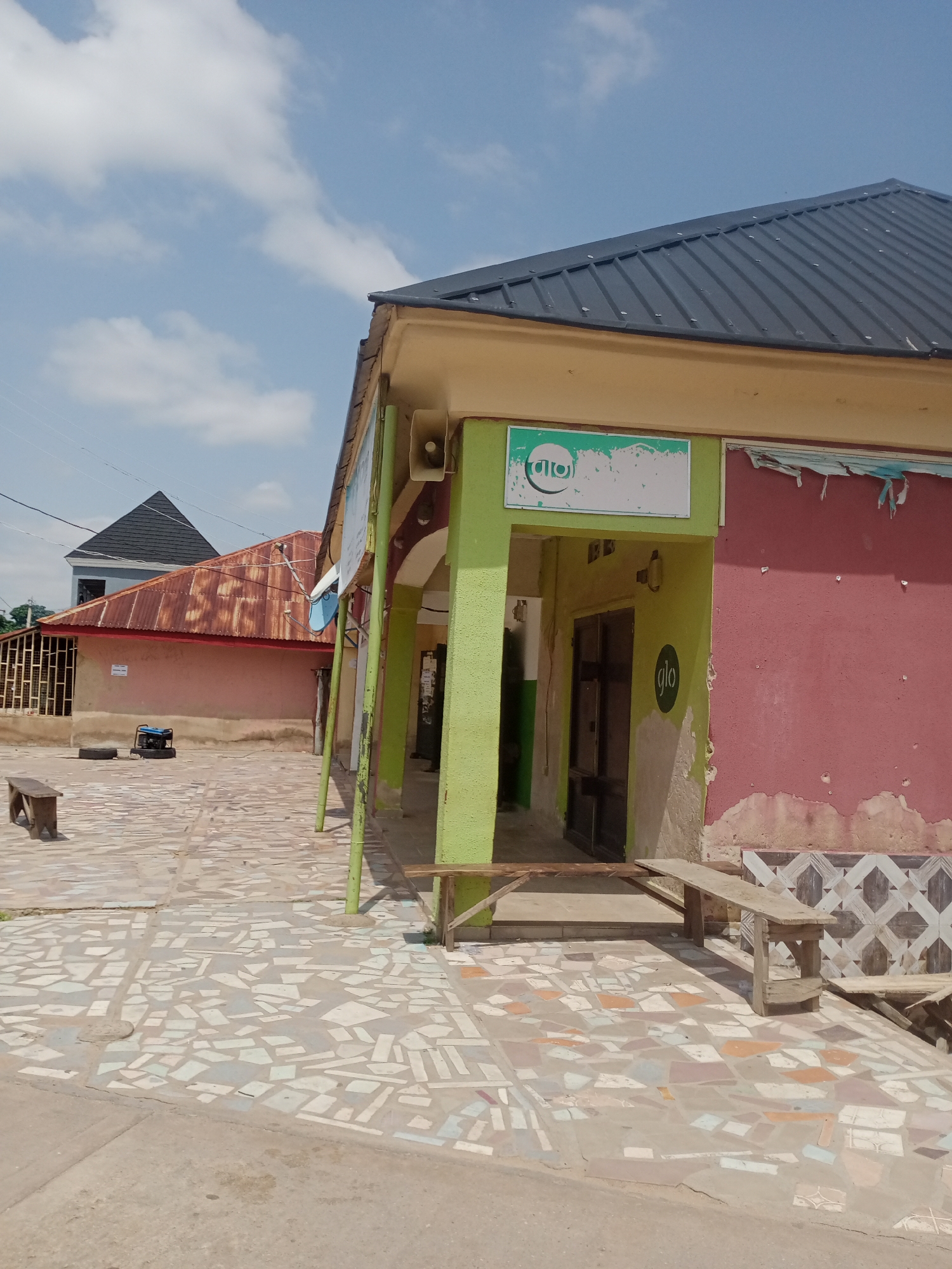 The image captures a storefront in Jalingo, Nigeria, under a partly cloudy sky. The building features a dark, corrugated metal roof and a light yellow awning. The front of the building is painted in two distinct sections: a vibrant green to the left, and a faded red on the right, with decorative tile work at the bottom of the red section. Two speaker units are mounted on the green section of the building. A sign with a circular logo and white text is affixed above the entrance, and another circular logo is visible on the red wall. The entrance itself appears to be a dark-framed doorway.
The ground in front of the building is paved with a mosaic of irregularly shaped ceramic tiles in various shades of white, gray, and light brown. Several wooden benches are placed on this tiled area. To the left of the building, a smaller building with a rusty, corrugated metal roof is visible, and behind it, a more modern building with a dark, pitched roof. A portable generator is also present on the tiled ground. The scene appears to be in a commercial or public area of the city.