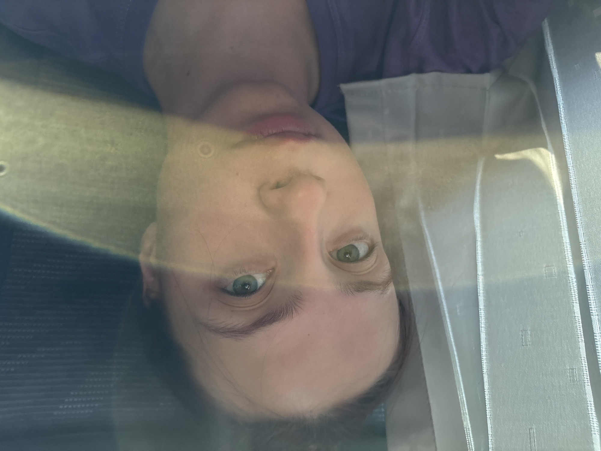 A person is seen through a glass window in Staryi Chul'tem, Russia. The person has light skin, and green eyes, and is wearing a purple top. The scene is bright. White curtains can be seen to the side of the person. A structured patterned object is on the bottom left.
